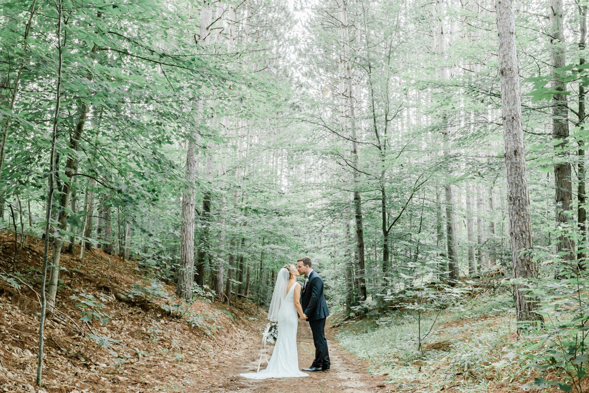 A couple hand in hand kissing in the middle of a forest in Ontario during the summertime for their wedding day