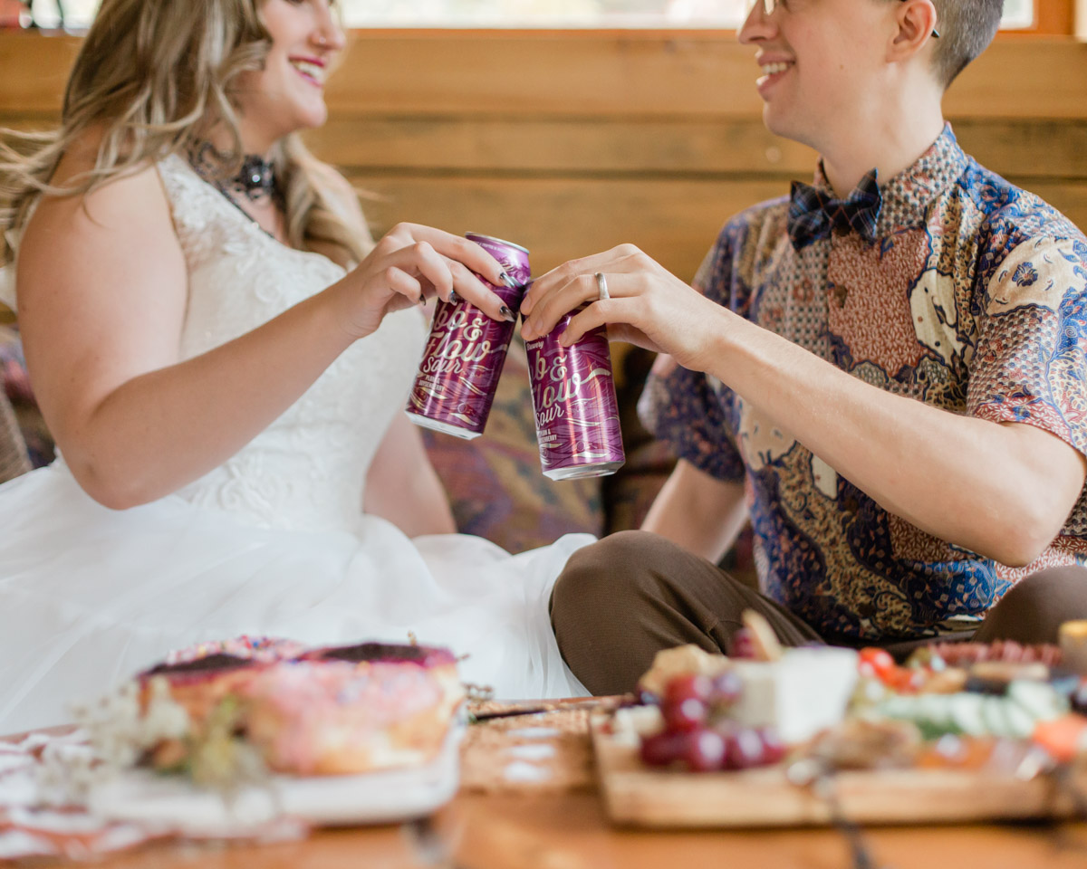 A couple enjoying their cozy cabin elopement in Muskoka Ontario cheering with their craft brew beer and enjoying a charcuterie board while laughing together. What a great wedding day! 