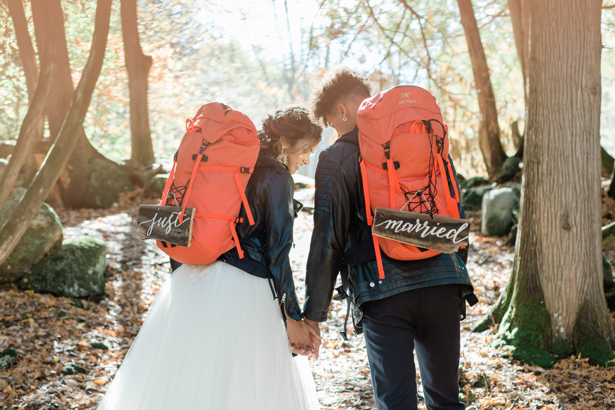 A couple hand in hand about to hike during their Ontario fall elopement. They have backpacks on that say just married and the sun is shining in front of them.