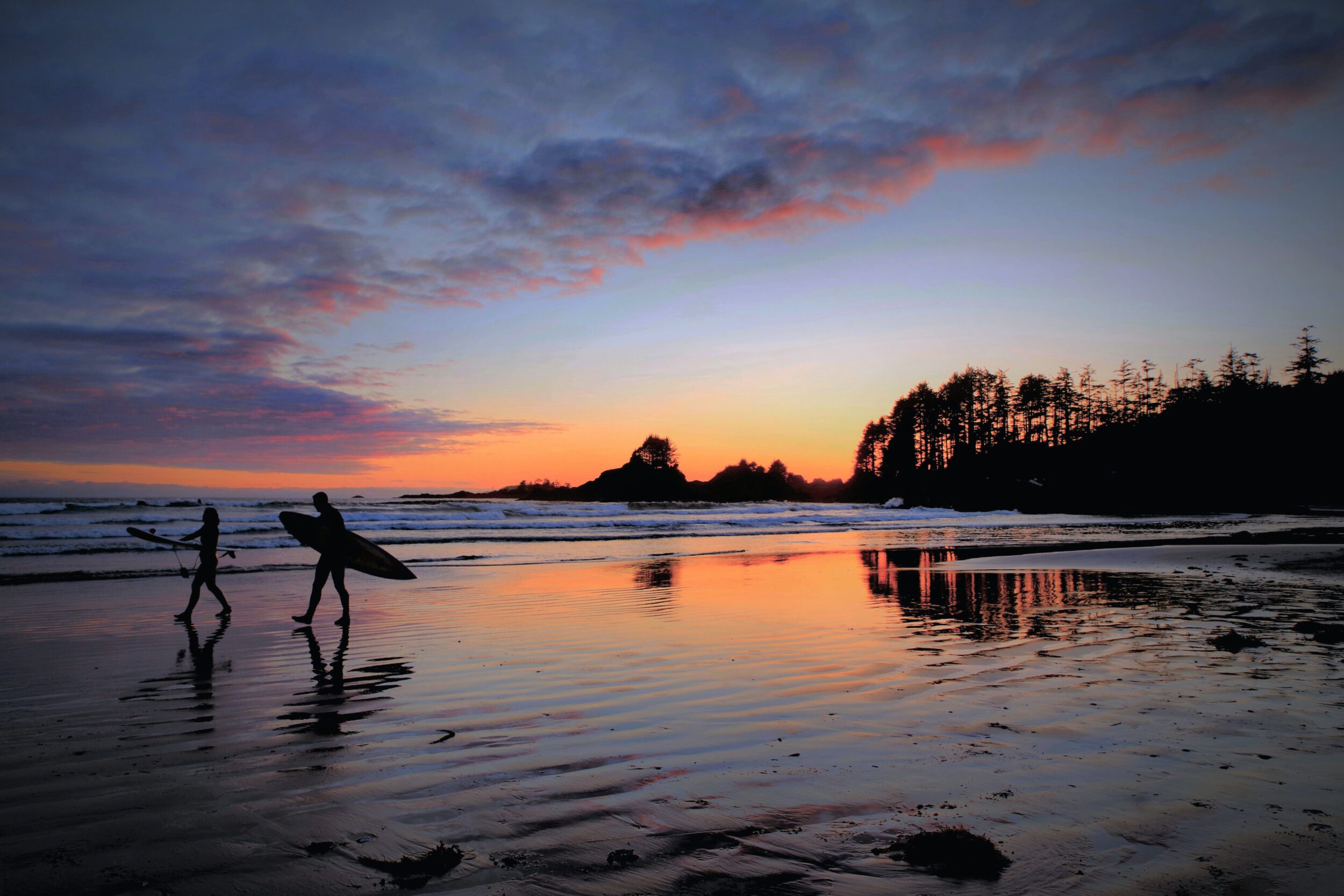 Surfers are photographed at Sunset walking along a beach in Tofino.