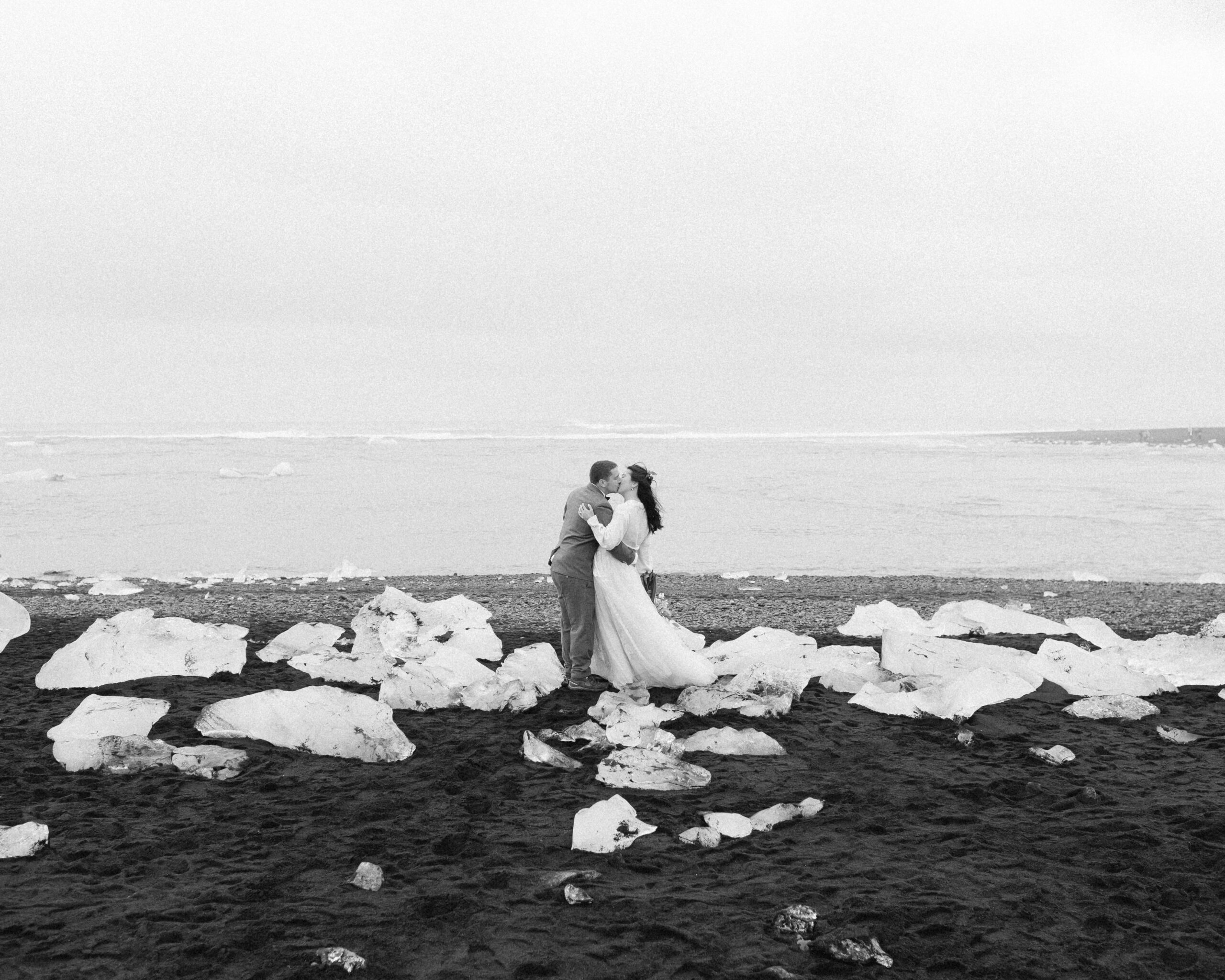 A couple is seen holding one another in wedding attire on an Icelandic black sand beach.