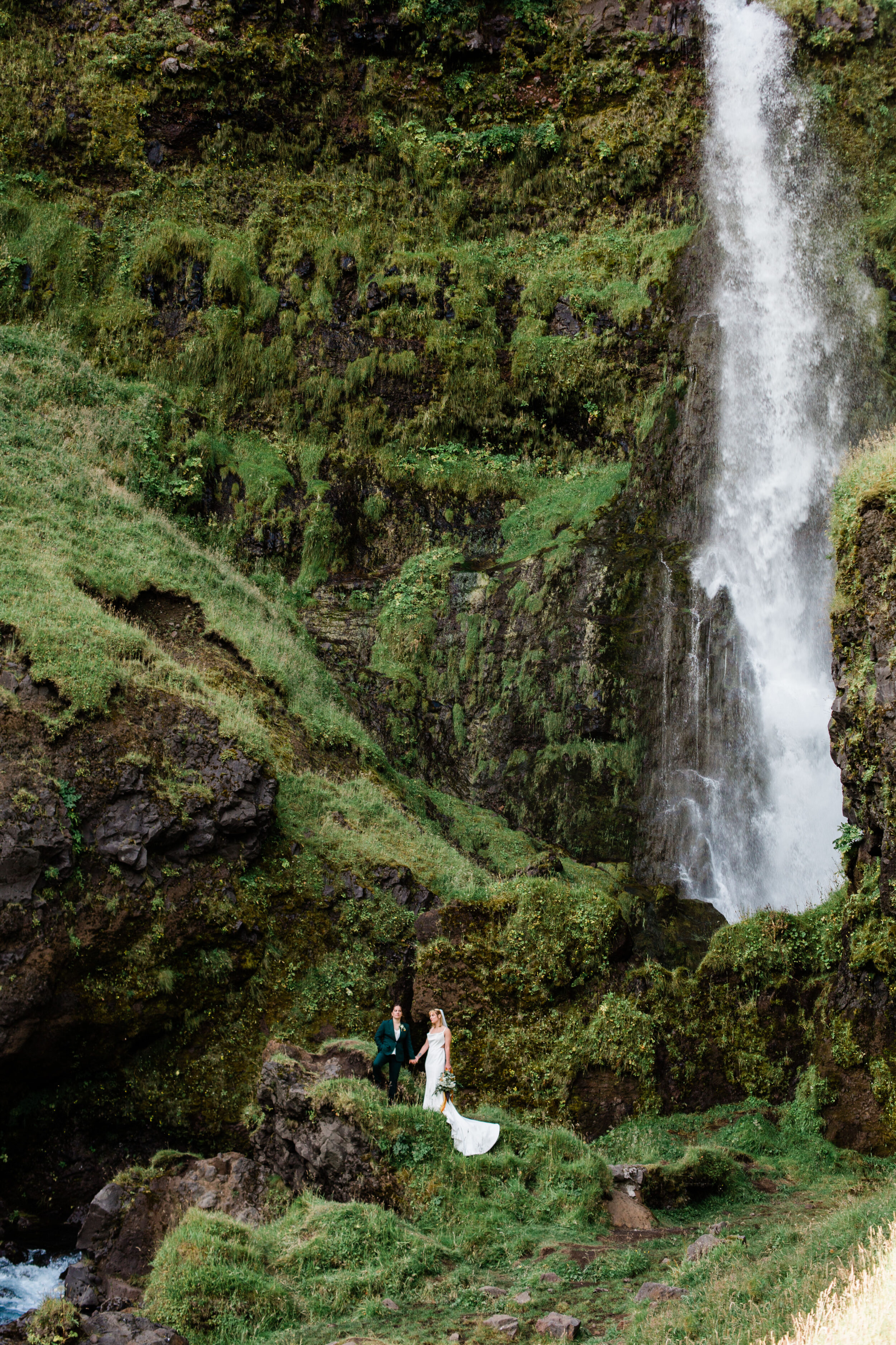 A couple stands amongst green cliffs by an Icelandic waterfall.