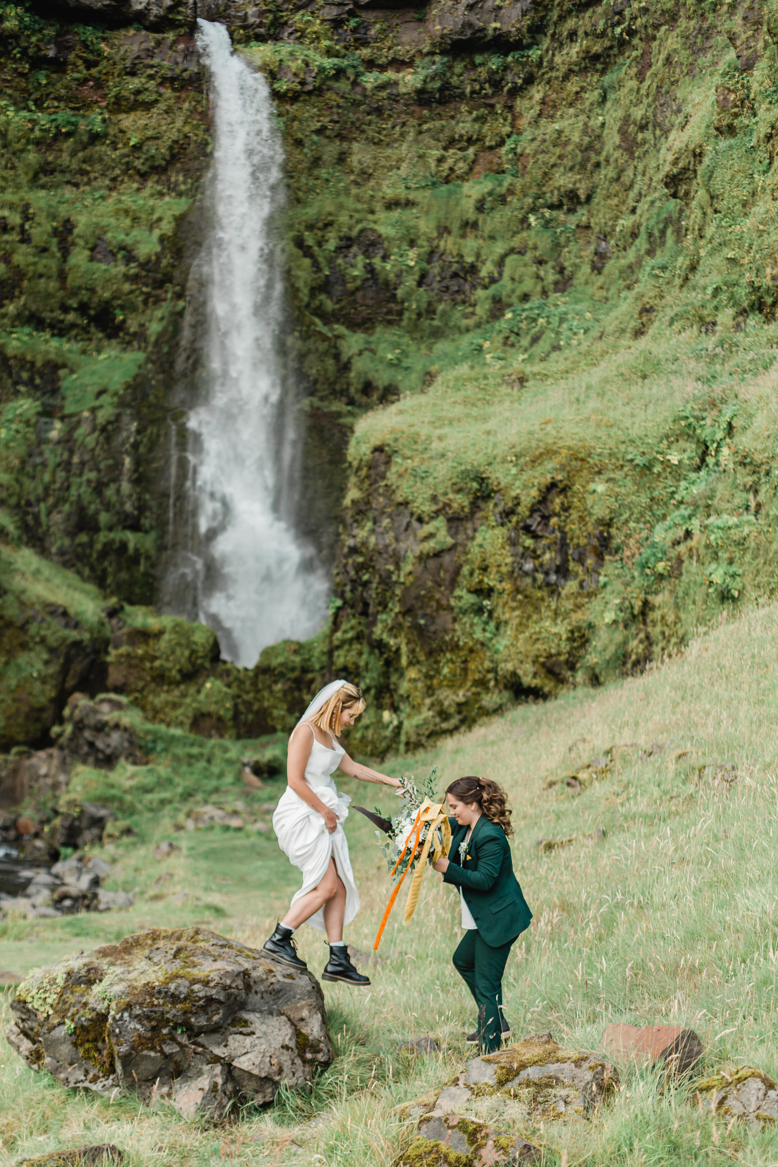 A woman helps her bride down from a rock while adventuring in Iceland during their wedding.