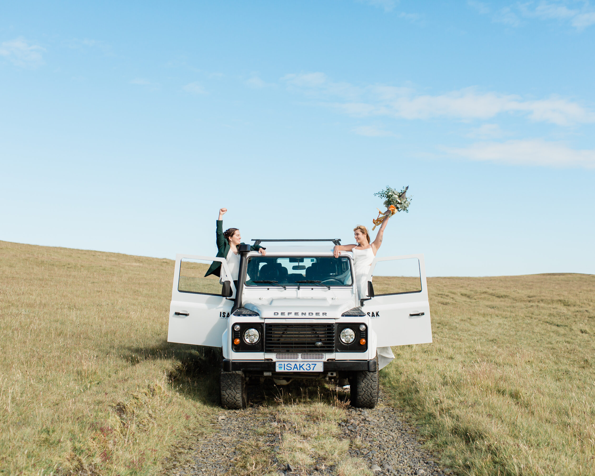 A couple cheers while hanging outside their white Land Rover getaway car.
