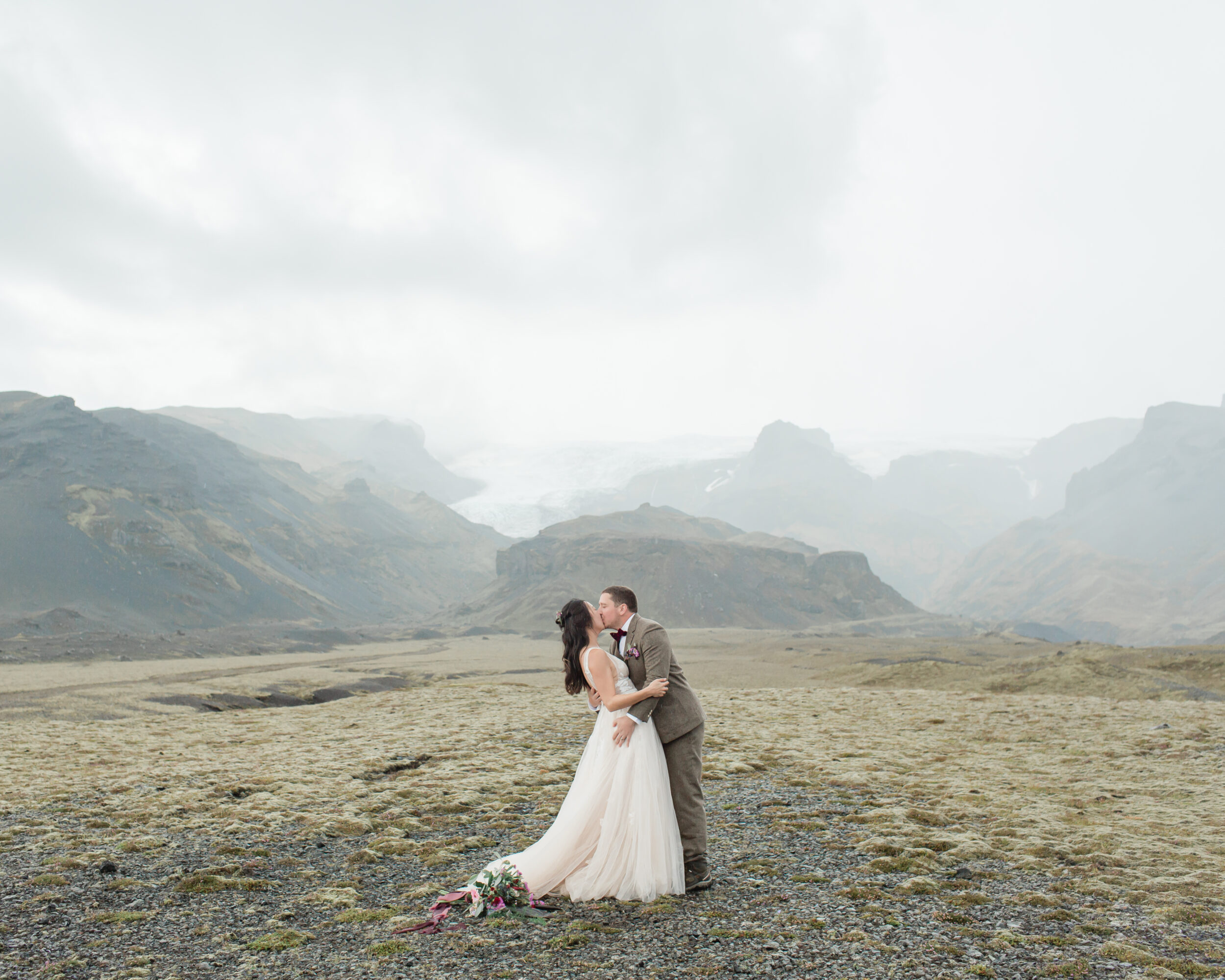 A couple in neutral wedding attire kisses beneath mountain peaks in Iceland.