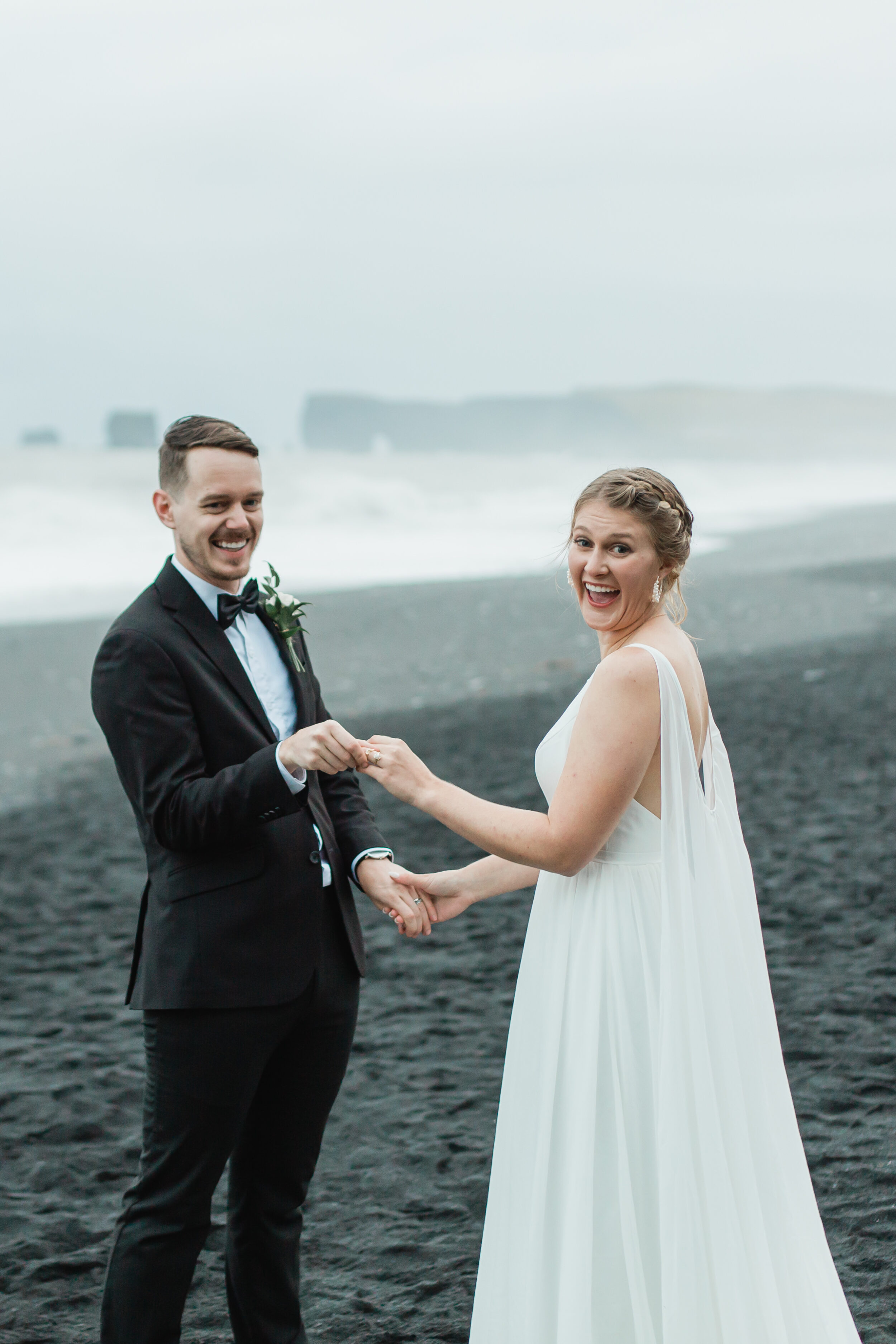 A couple smiles at the camera while holding hands on a black sand beach in Iceland.
