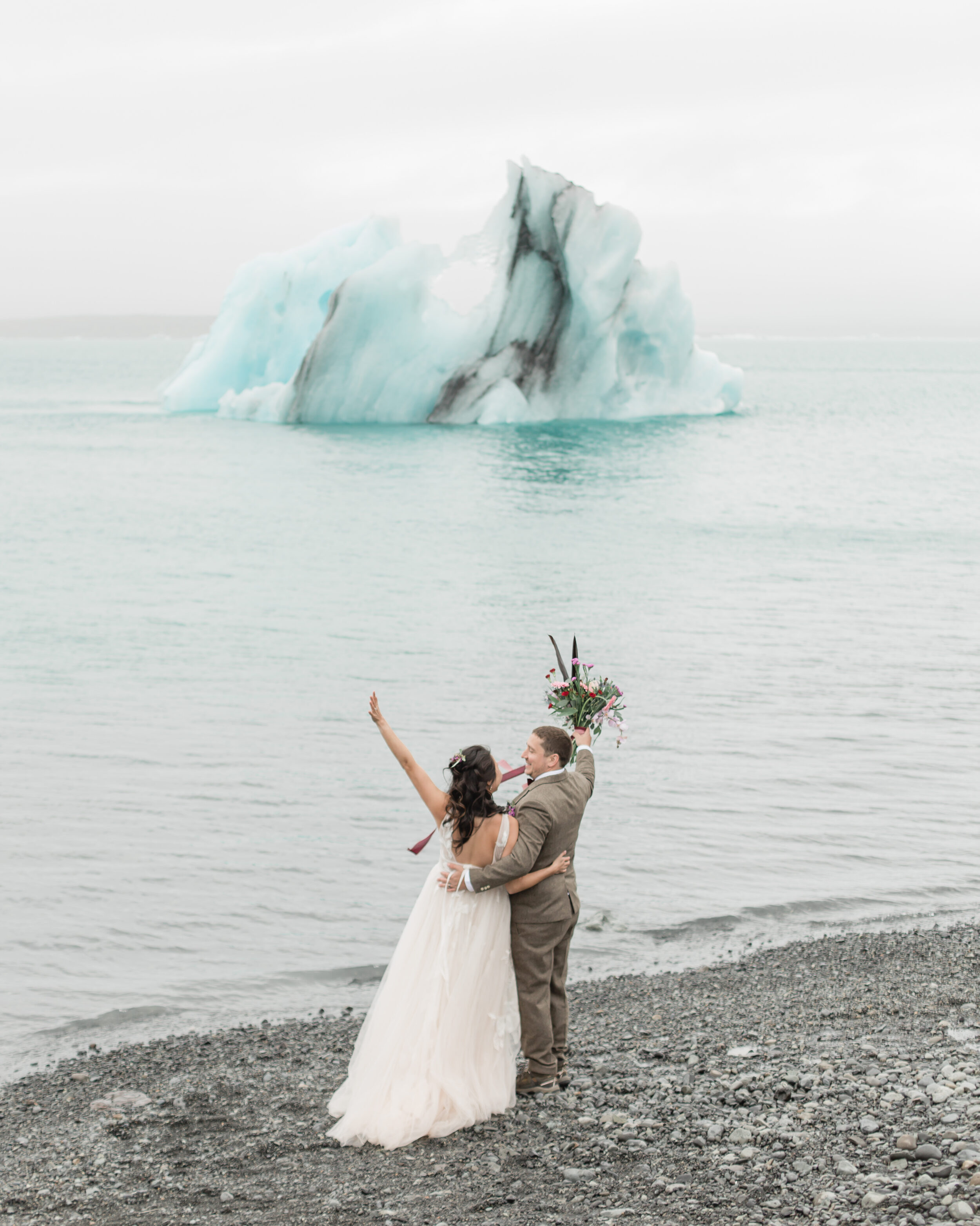 A couple cheers on a beach during their Iceland with an iceberg in the distance.