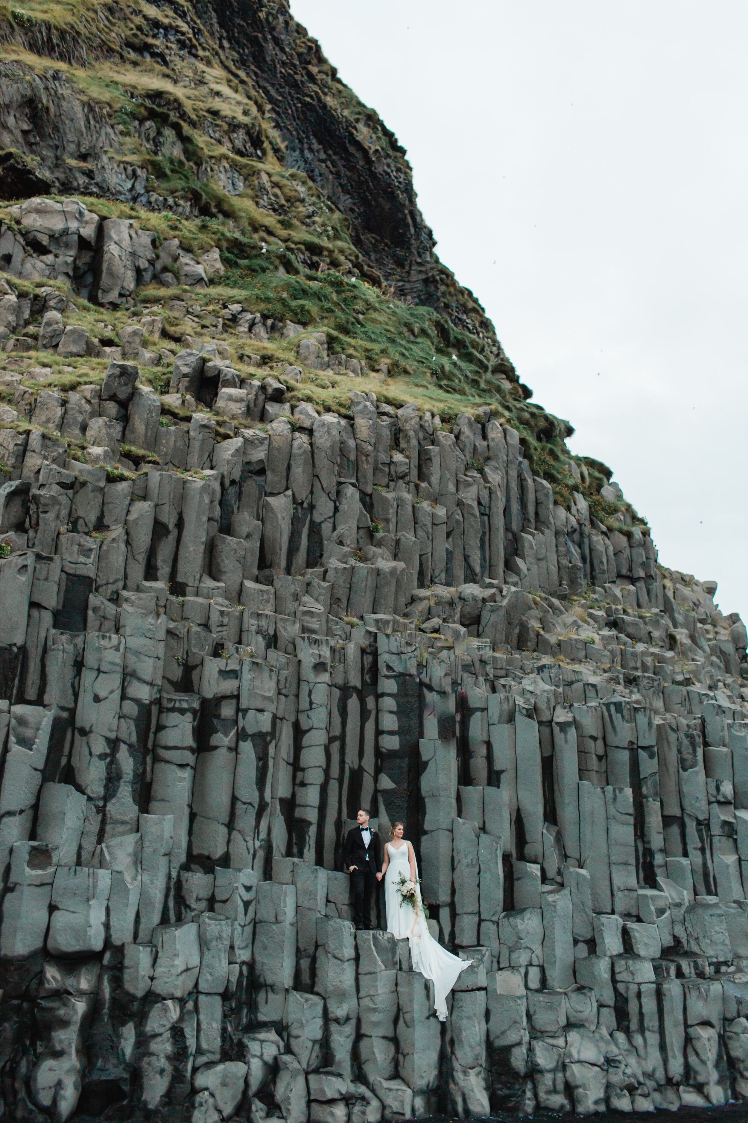 Newlyweds stand on large basalt columns in Iceland near a black sand beach.