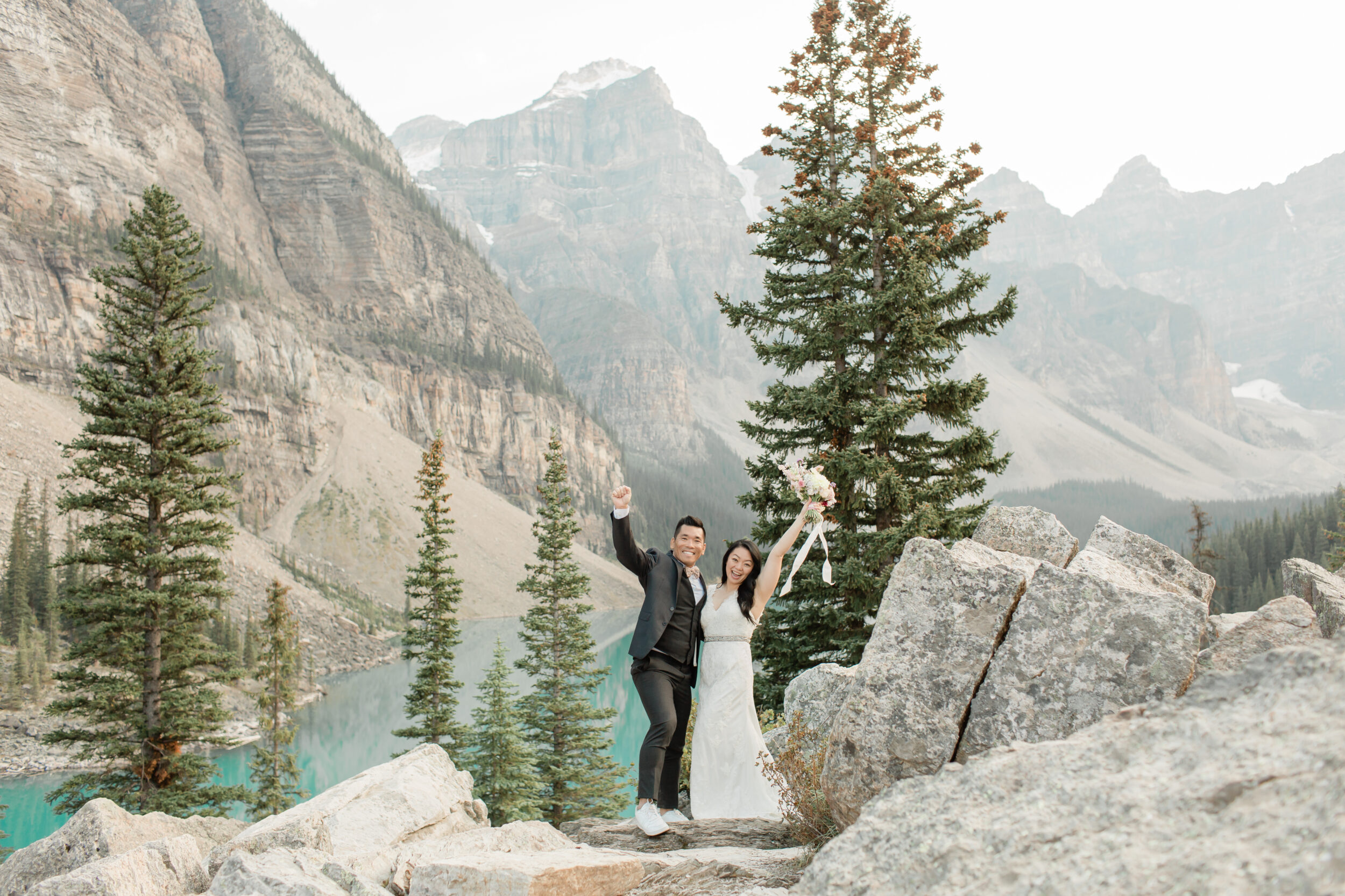 A couple cheers while celebrating their Banff elopement near evergreens and a lake in the Canadian Rockies. 