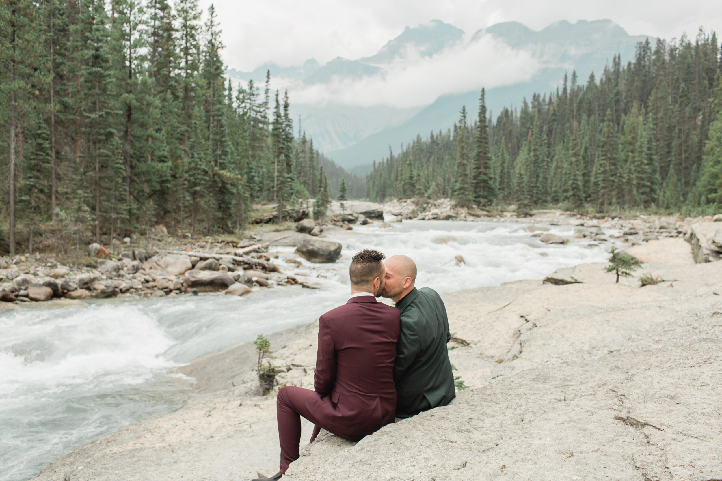 A couple shares a kiss near a river and mountainside in Banff.