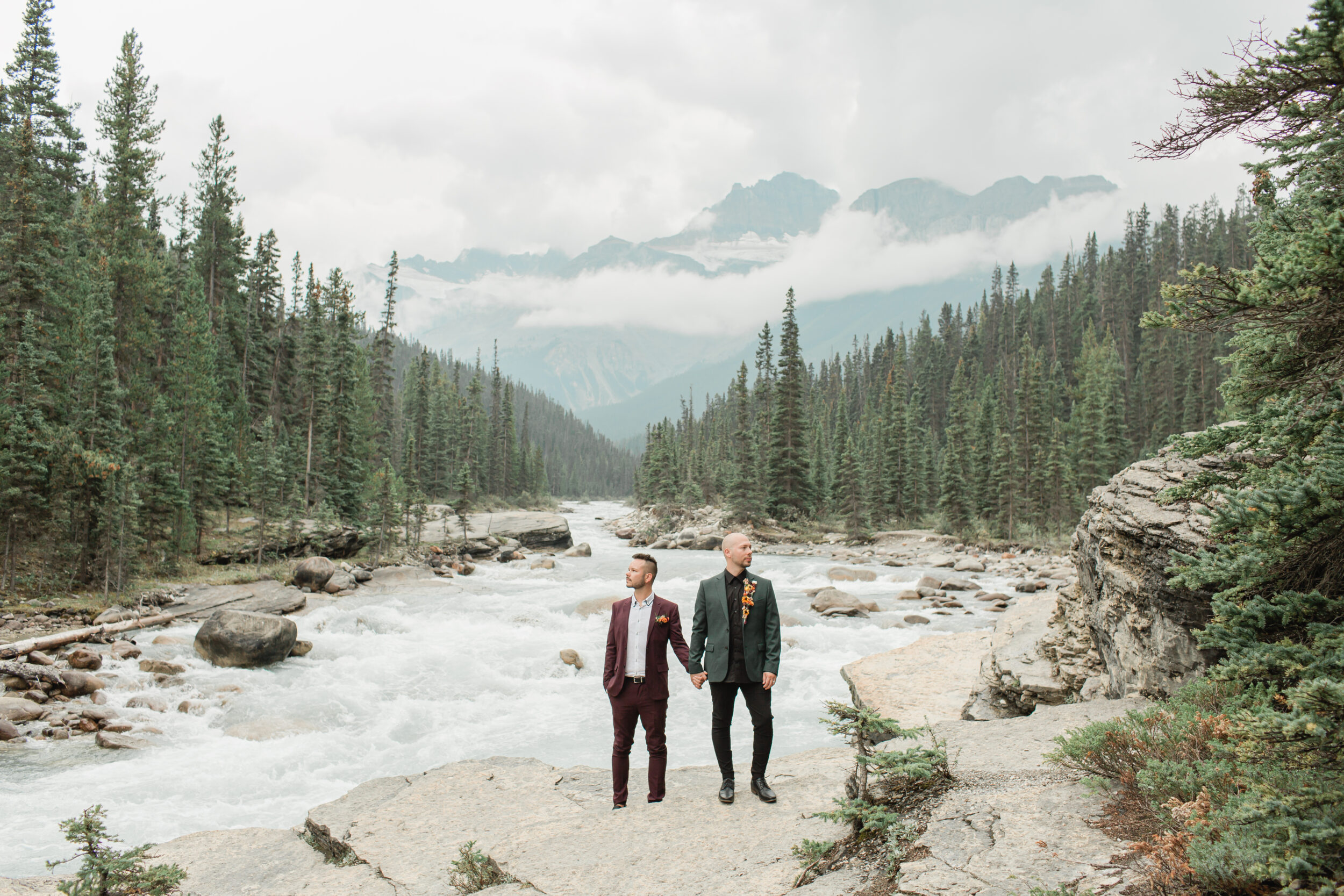 A couple holds hands on a river's edge during their intimate adventure elopement in Alberta.