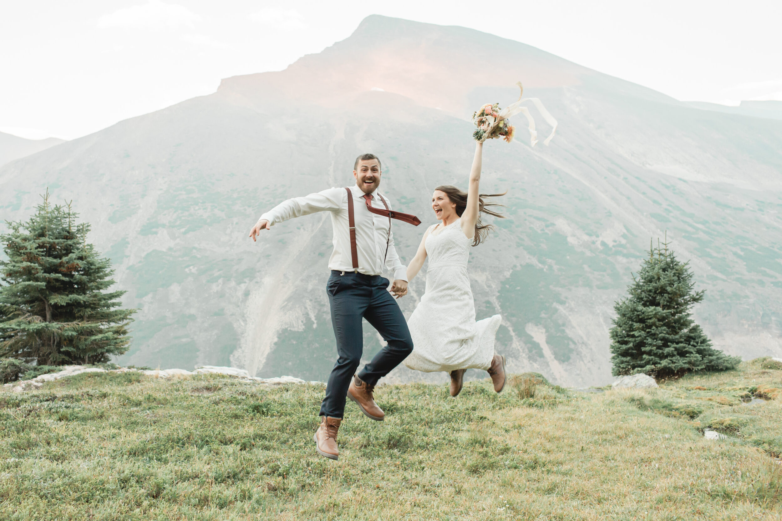 A couple is seen jumping in excitement on a green mountainside after their Banff elopement.