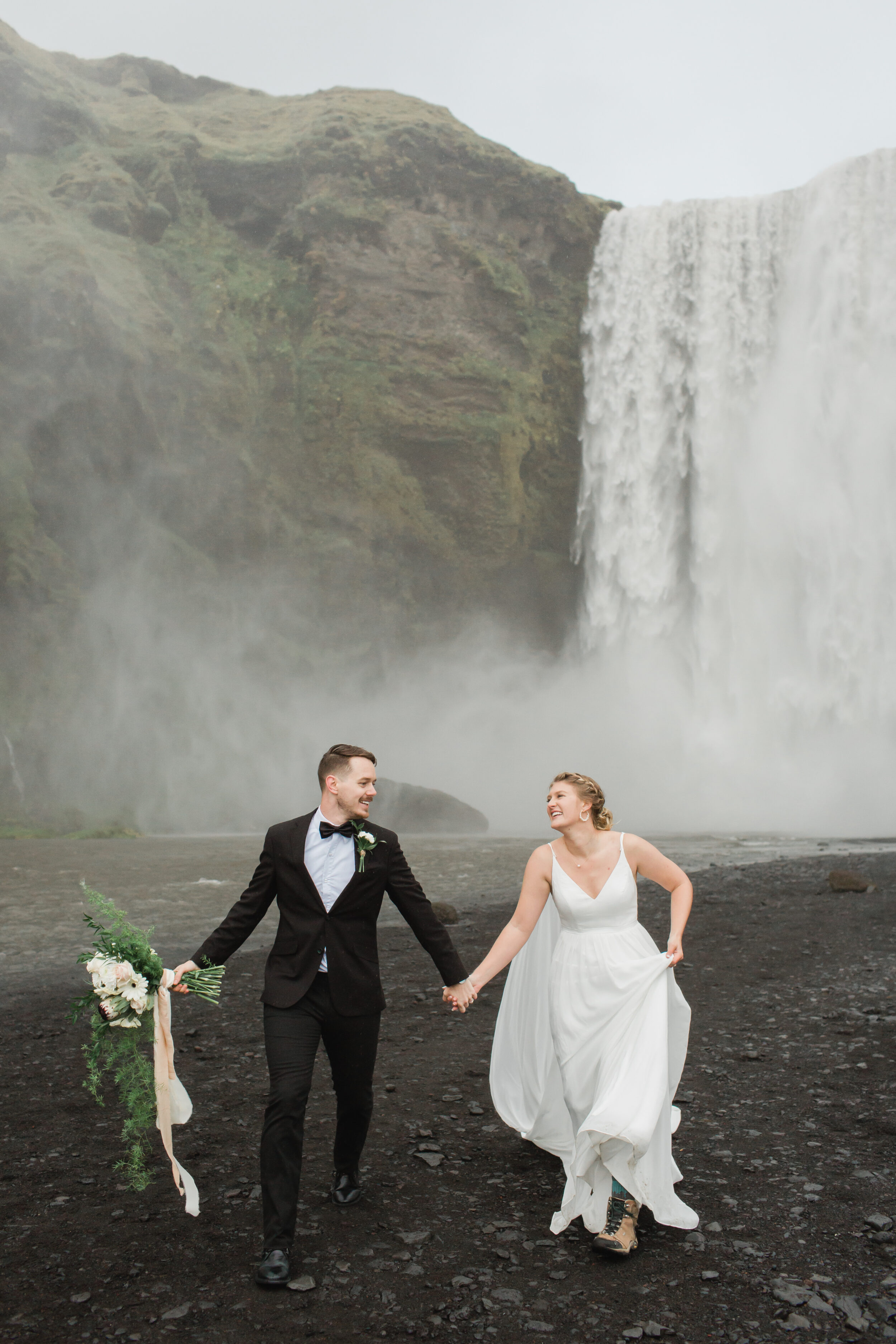 A man holds his bride's wedding florals as they run away from Skogafoss.