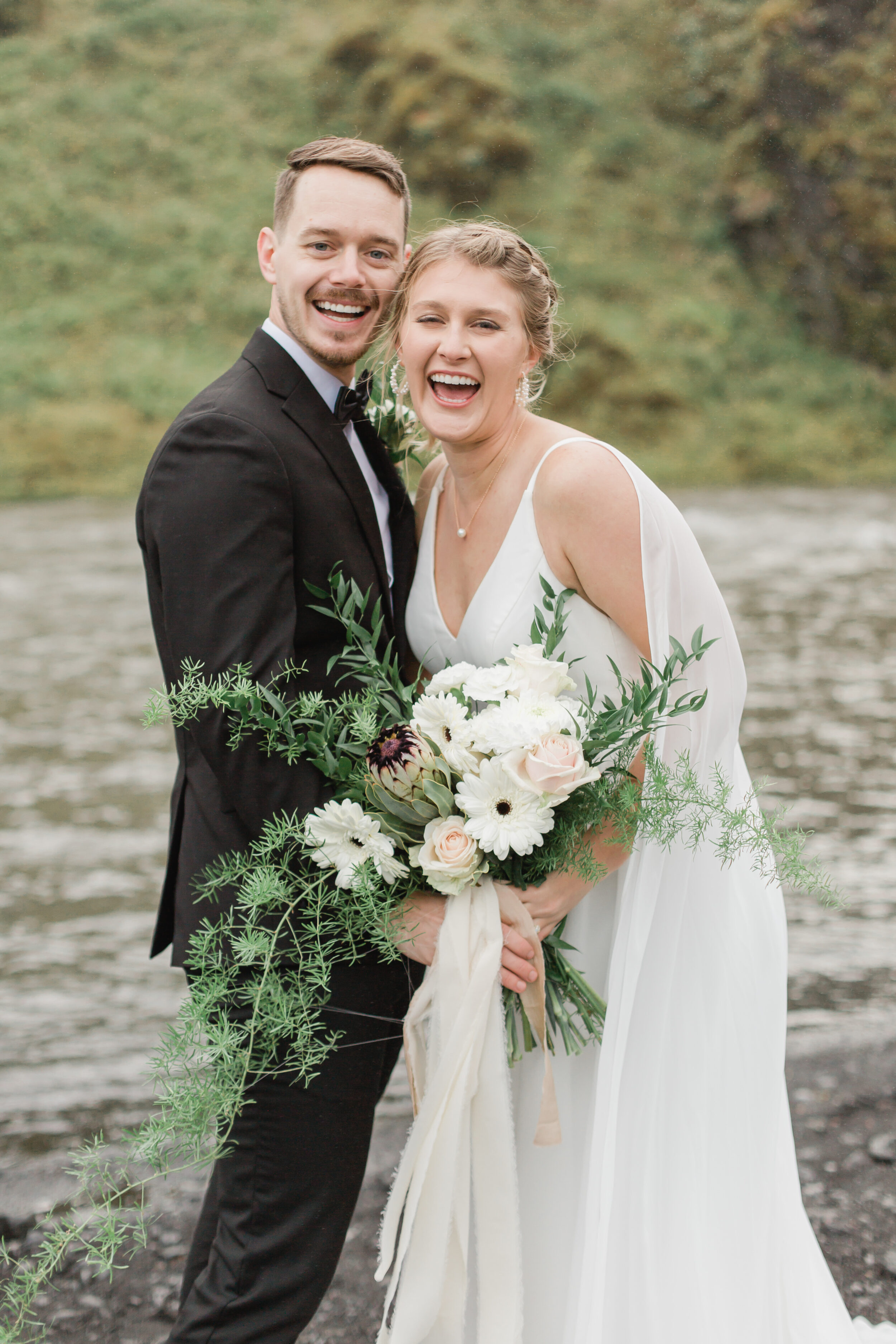 A couple smiles at the camera and holds large wedding florals in Iceland. 