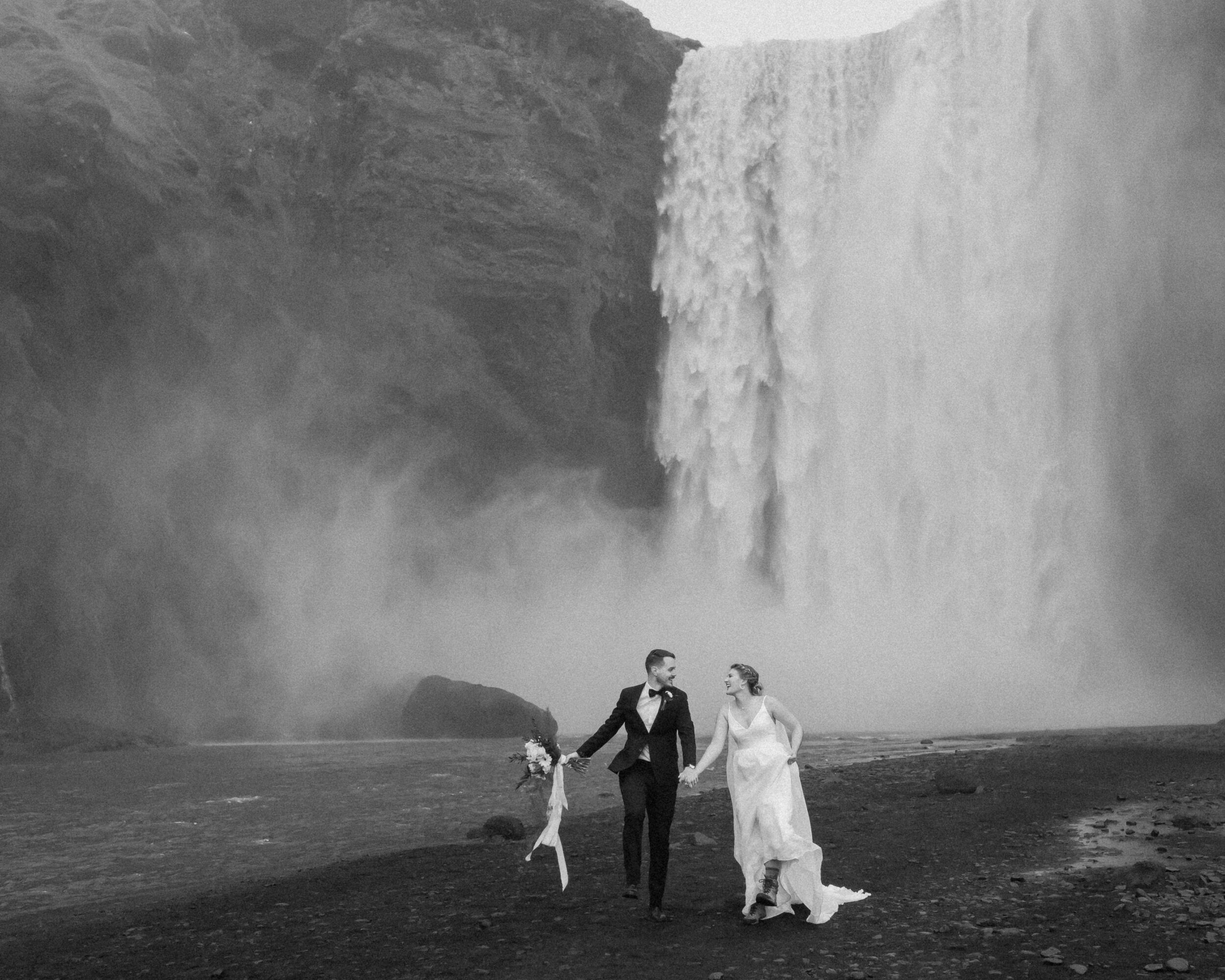 A couple walks together beneath an Icelandic waterfall. 