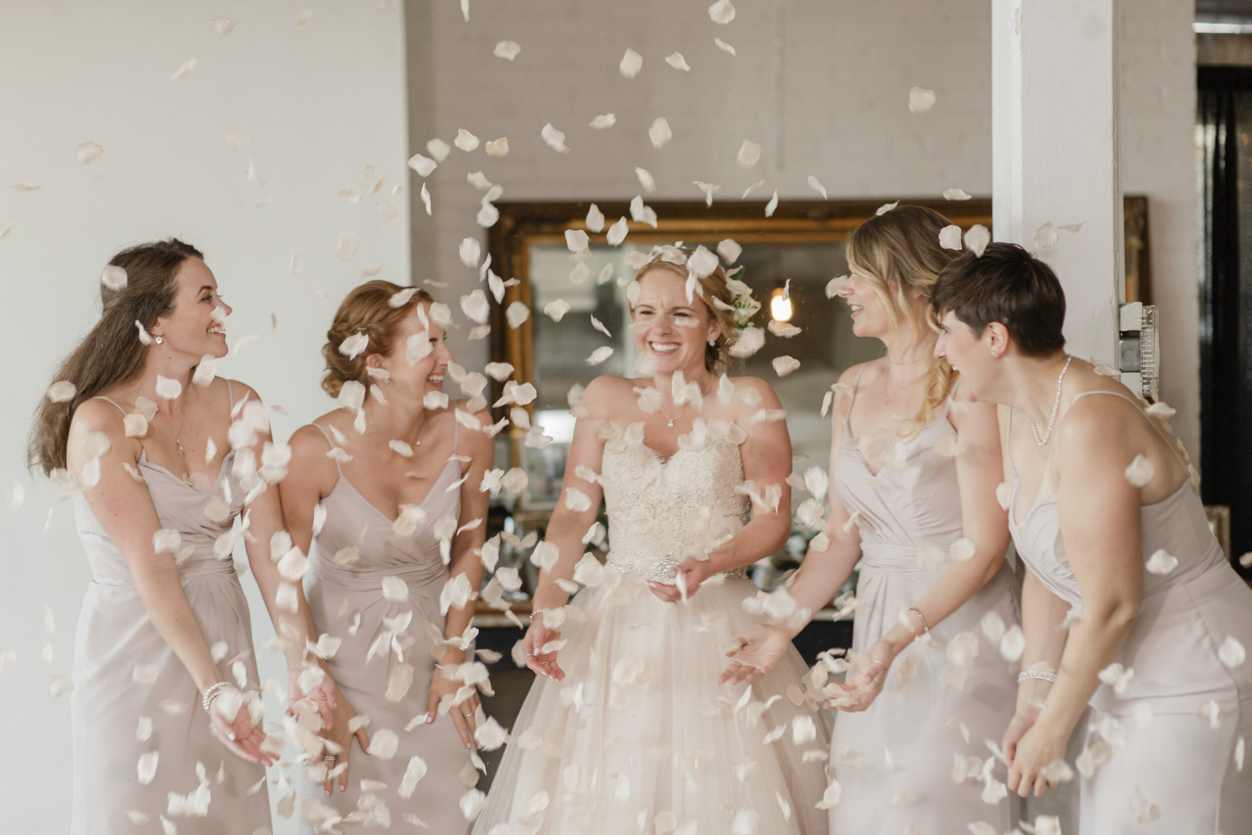 Bridesmaids toss flower petals as a bride enters a white room. 