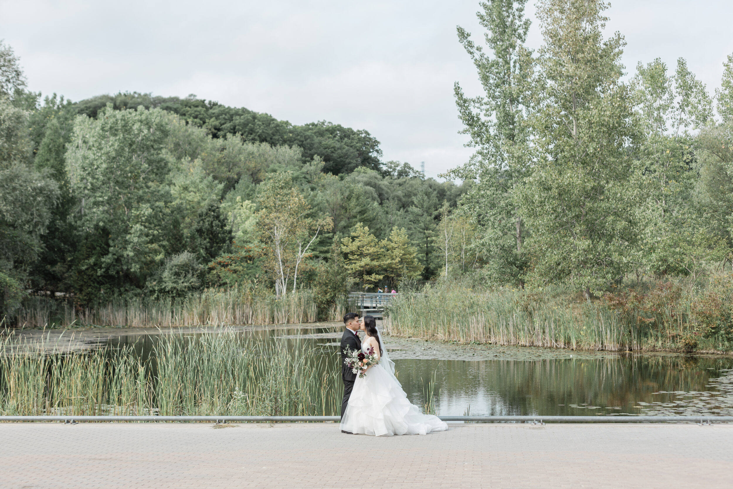 A couple in wedding attire kisses in front of a pond in Toronto during their elopement.