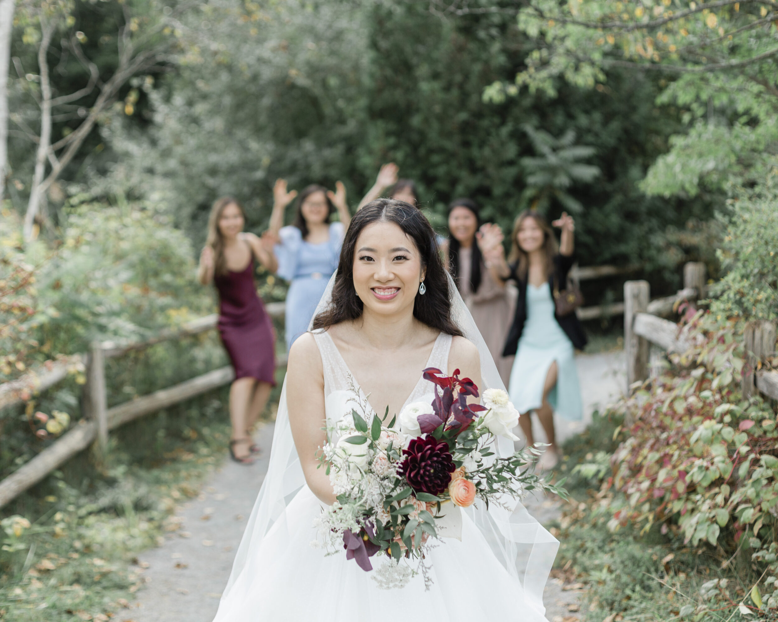 A bride walks down a Toronto nature path with her bridal party celebrating behind her.