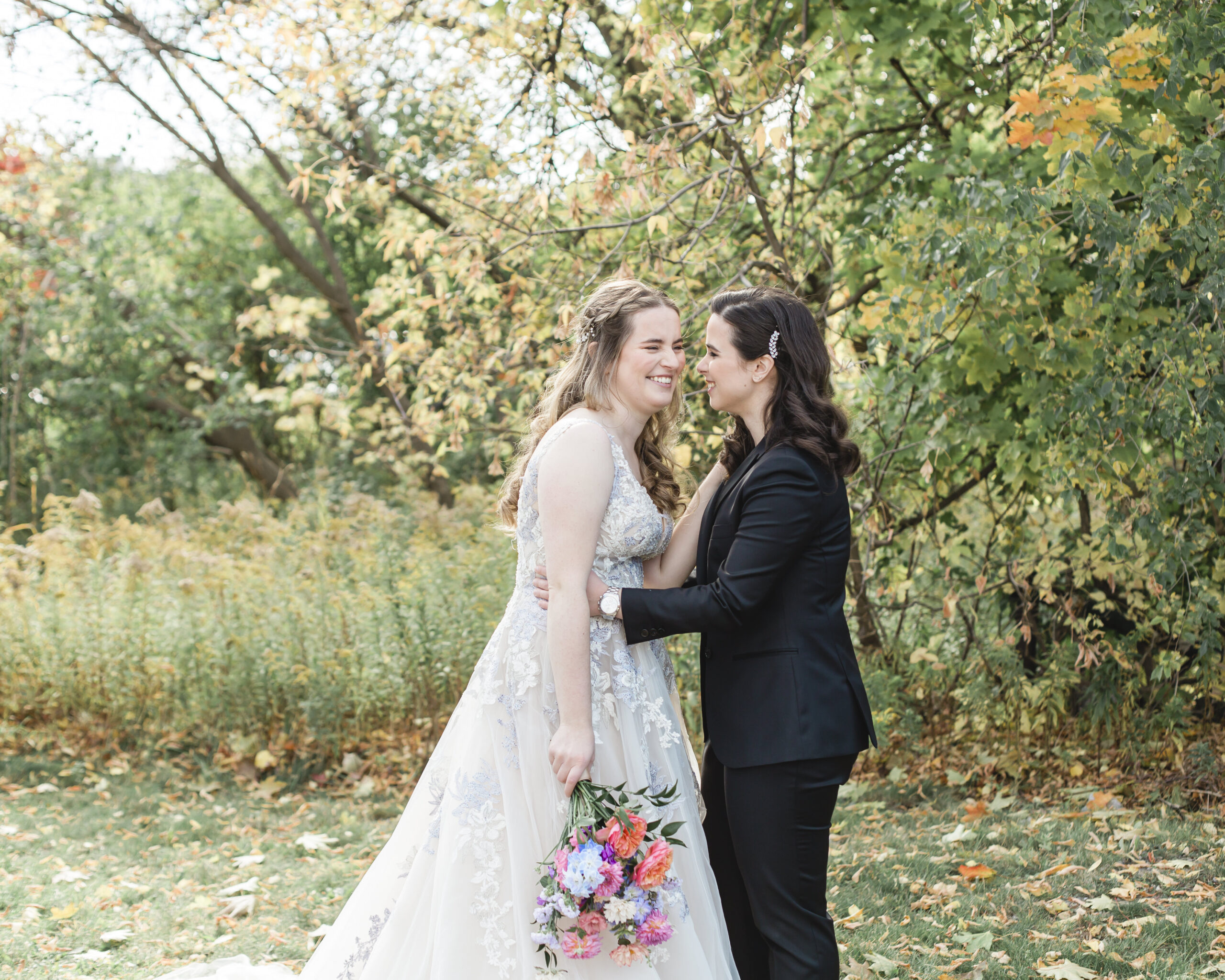 Two brides hold one another while holding a colorful floral bouquet. 
