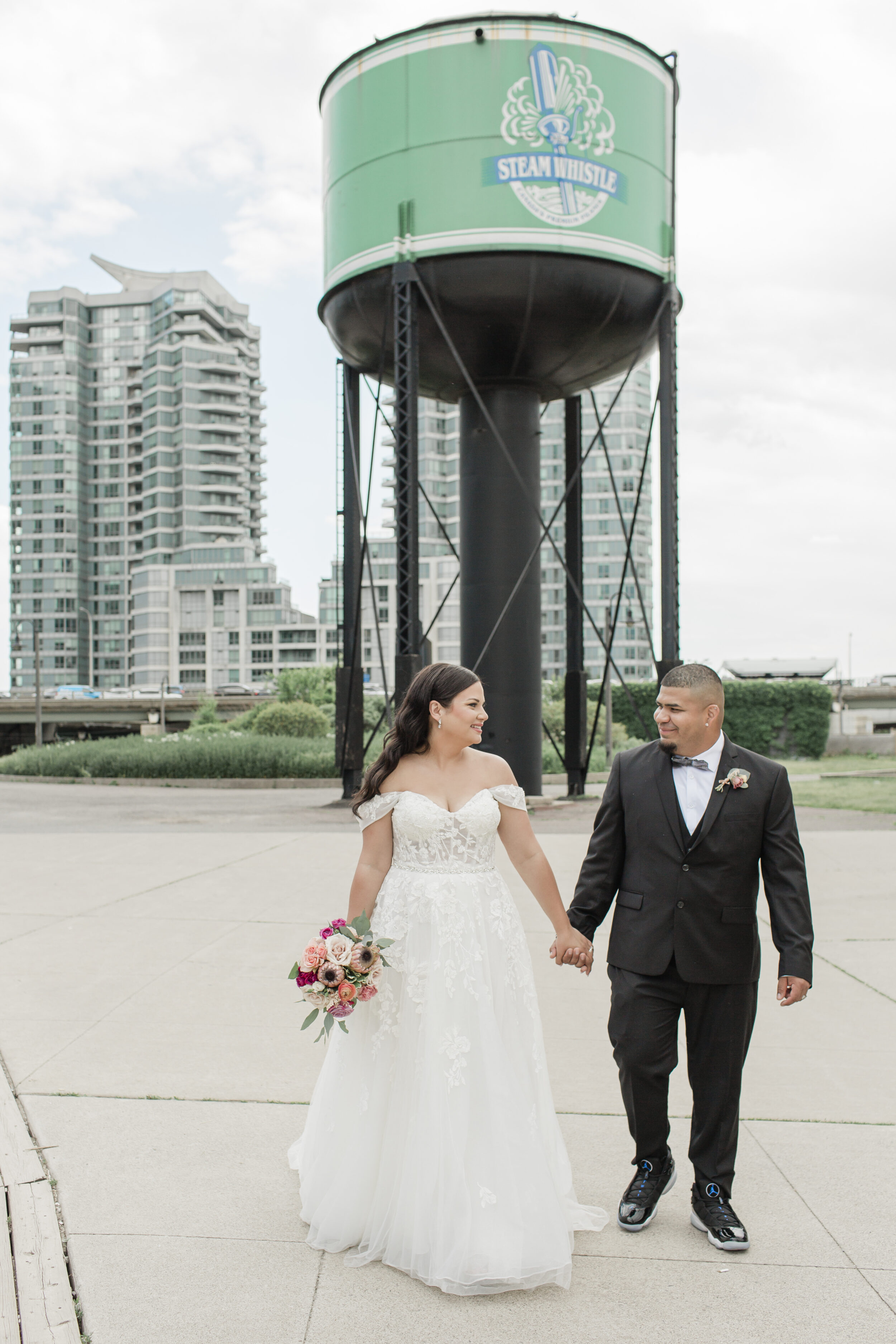 A couple stands beneath a water tower in downtown Toronto while wearing wedding attire.