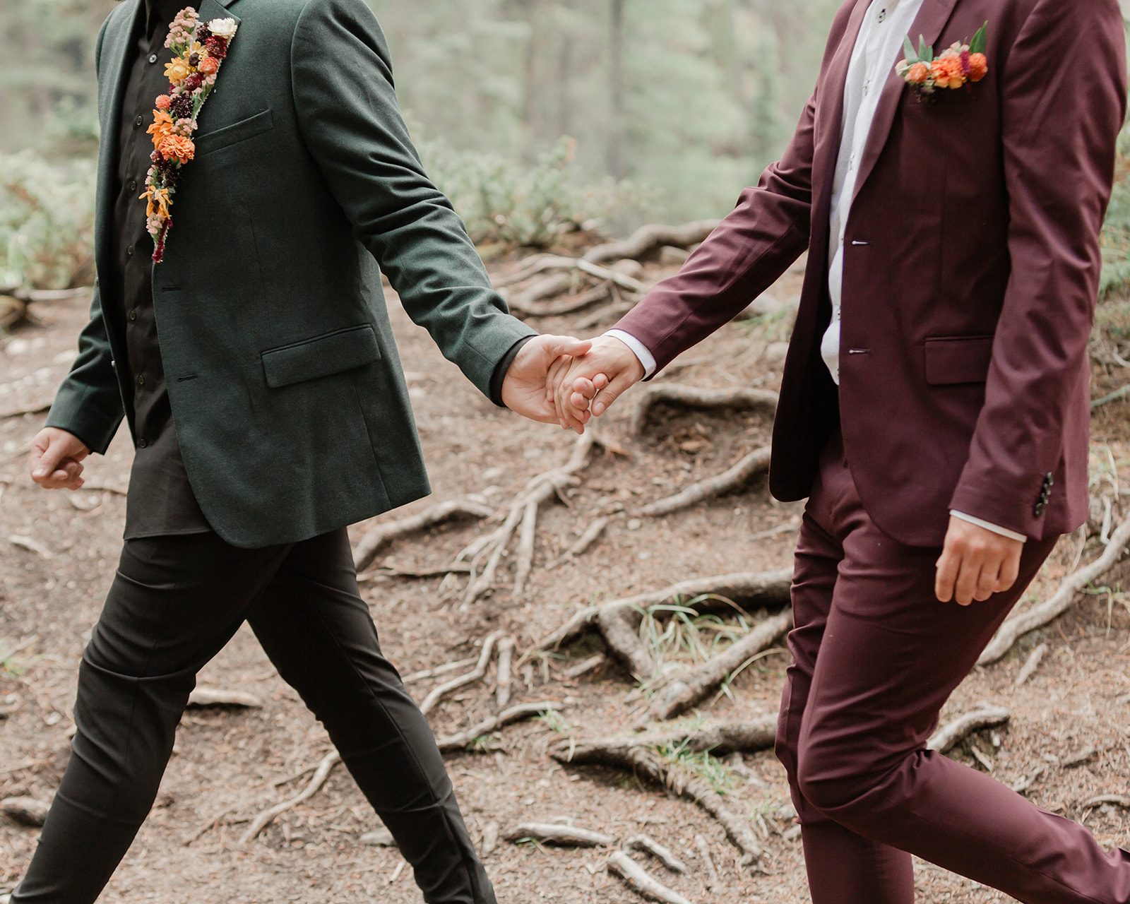 A man in a forest green suit leads his groom, who is wearing a burgundy suit, through Mistaya Canyon during their elopement.