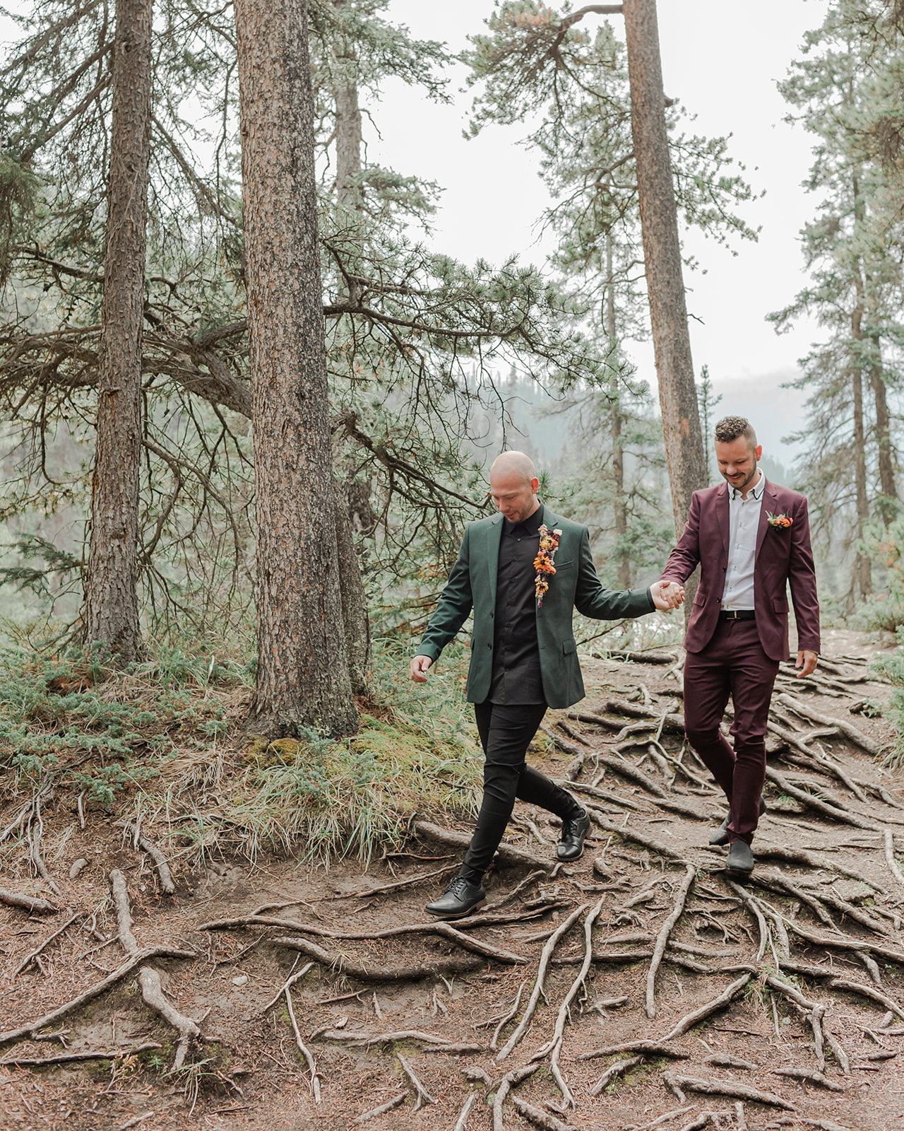 A man in a forest green suit leads his groom, who is wearing a burgundy suit, through Mistaya Canyon during their elopement.