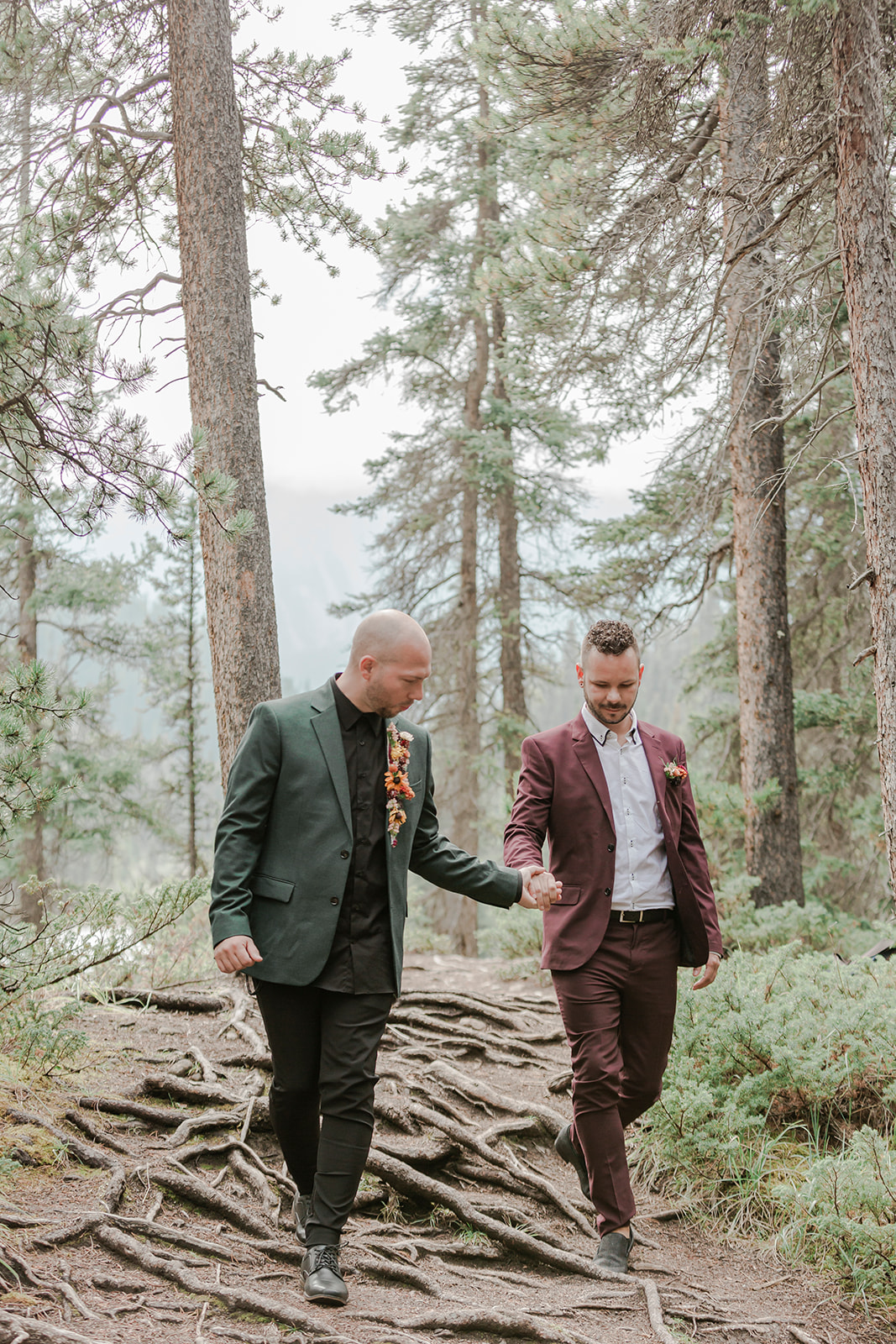 A man in a forest green suit leads his groom, who is wearing a burgundy suit, through Mistaya Canyon during their elopement.