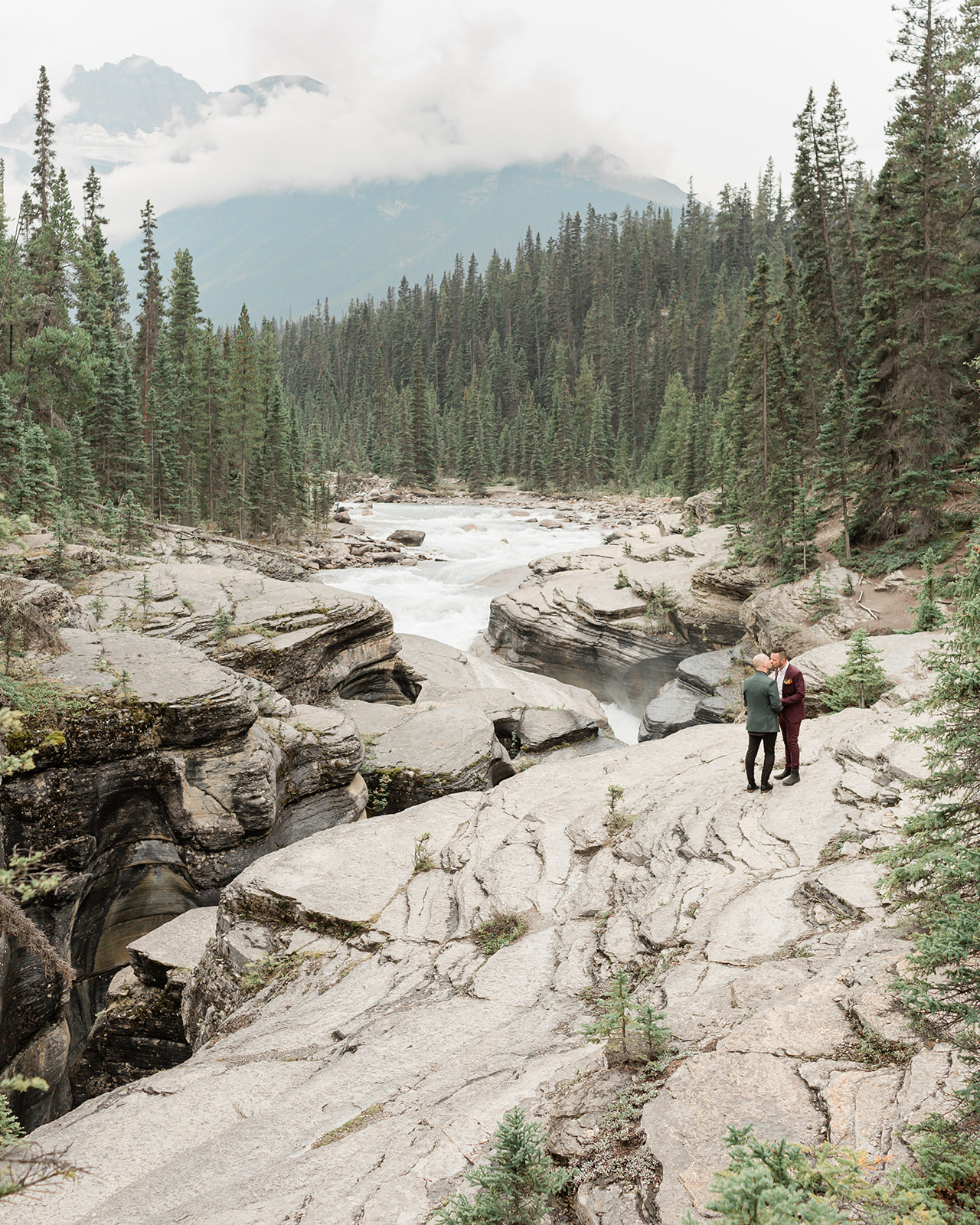 A man in a burgundy suit kisses his groom, who is wearing a forest green suit, while standing on a rock formation during their Mistaya Canyon elopement.