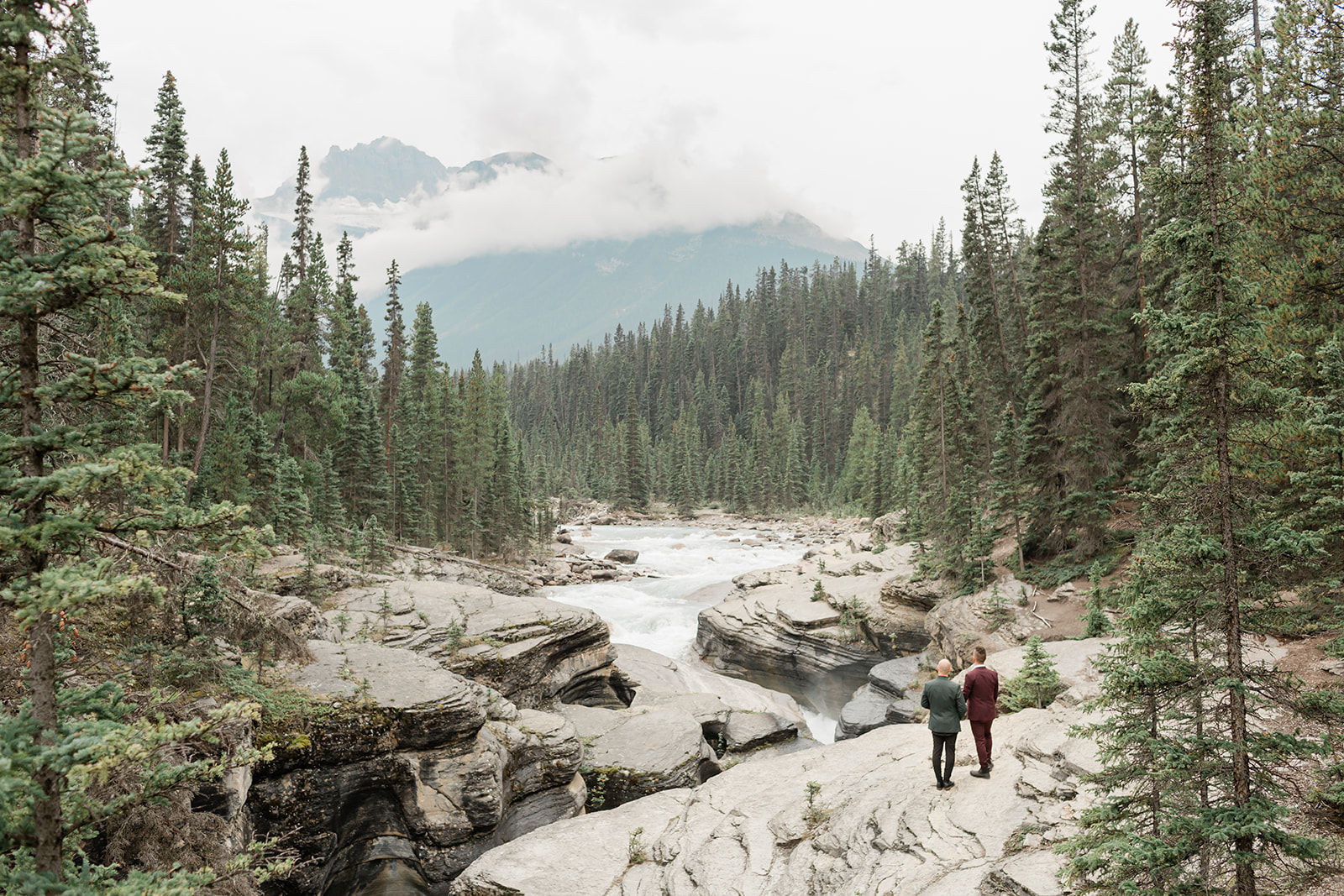 A Mistaya Canyon elopement couple holds hands while admiring a canyon and mountain in the distance. 