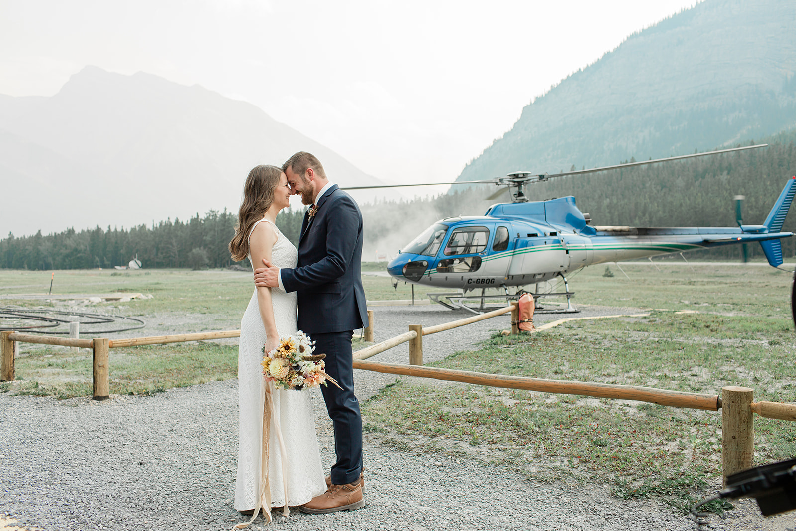 A couple in formal wedding attire embraces near a helicopter during their Banff helicopter wedding. 