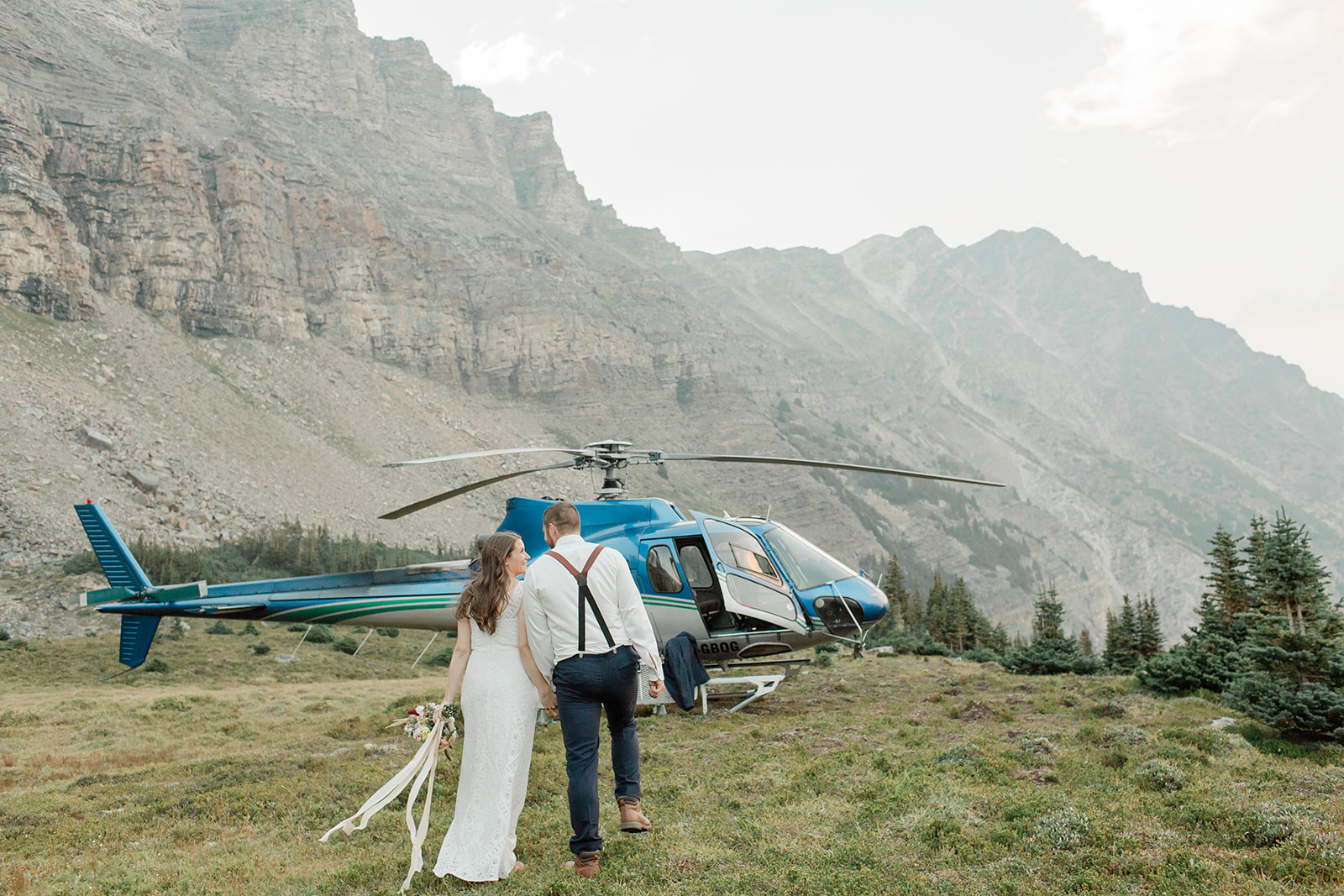 A couple in formal wedding attire walks towards a Rockies Heli 
helicopter during their Banff helicopter wedding. 