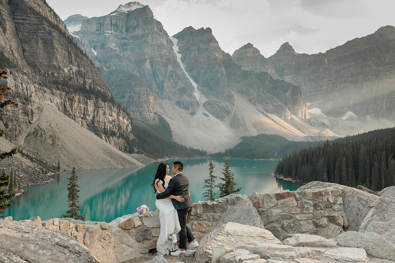 A Banff National Park elopement couple leans against a wall that overlooks an alpine lake. 