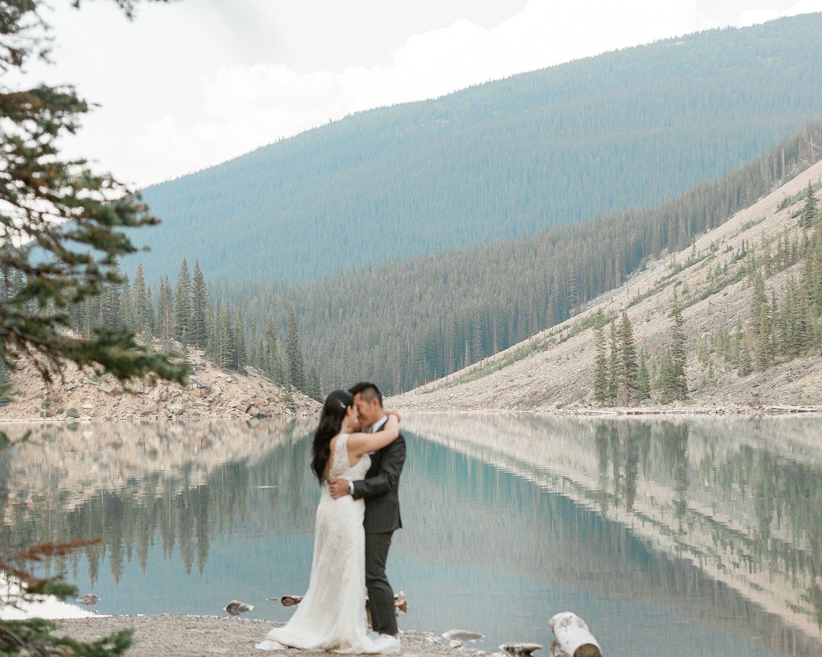 An adventure elopement couple in formal wedding attire embraces while standing on the shores of a lake in Banff National Park. 