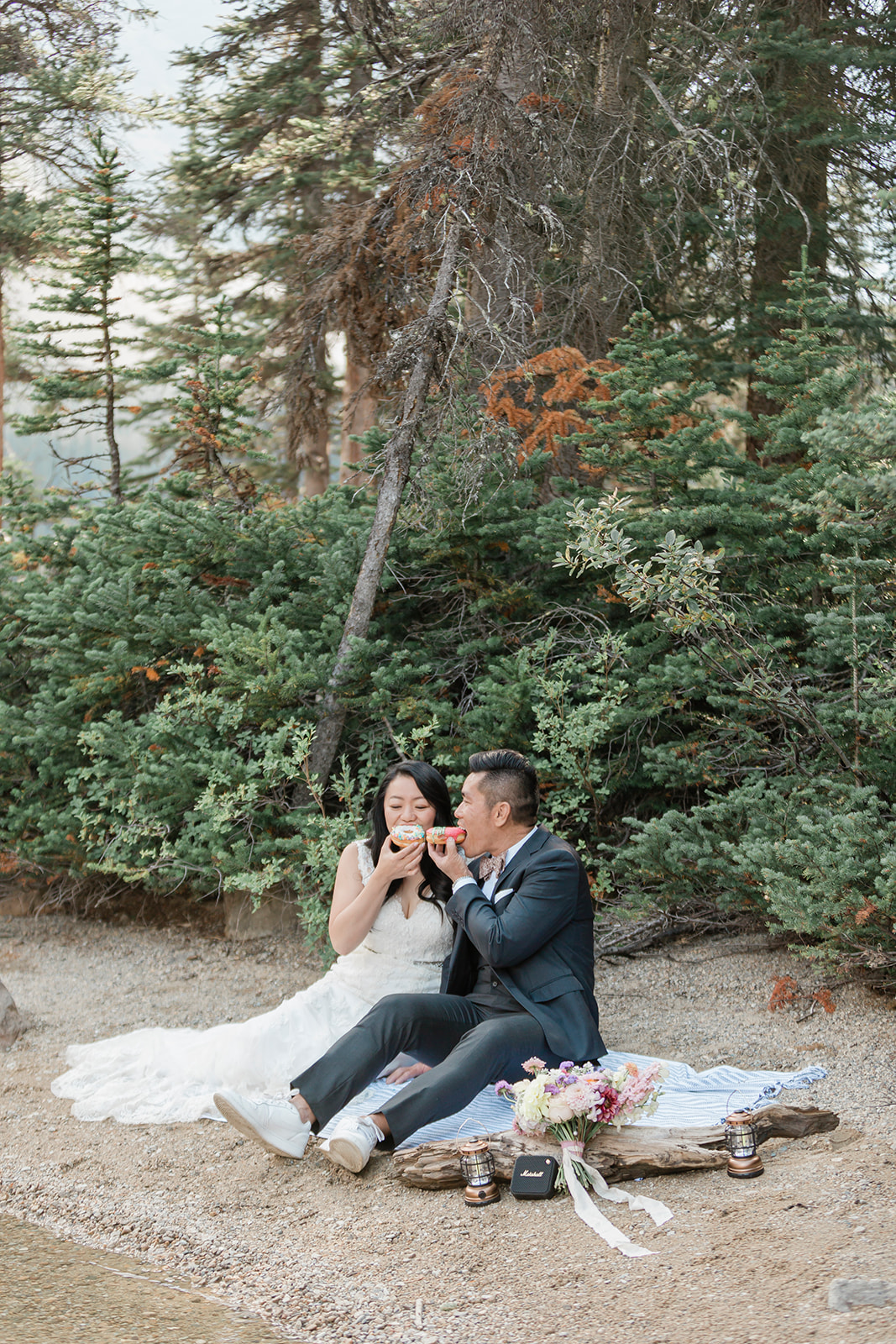 A couple in formal wedding attire shares a lakeside picnic with donuts in Banff National Park. 