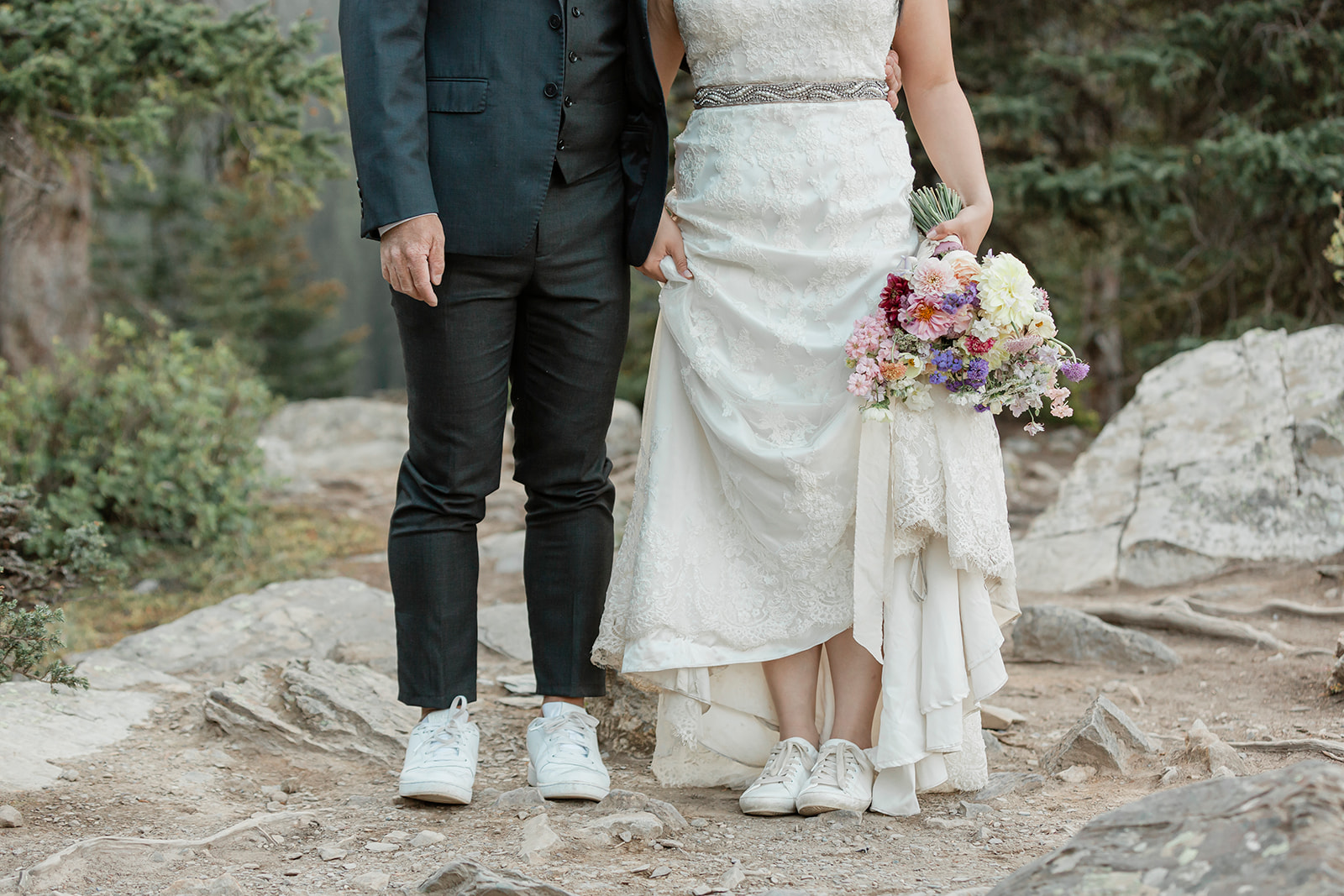 A bride in a white, lace wedding gown holds a bright, pastel bouquet and her groom's hand, who is wearing a suit and white shoes. 