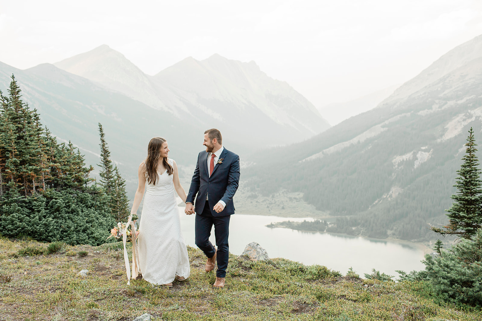 A Banff helicopter elopement couple holds hands while walking near an overlook near an alpine lake and mountains. 