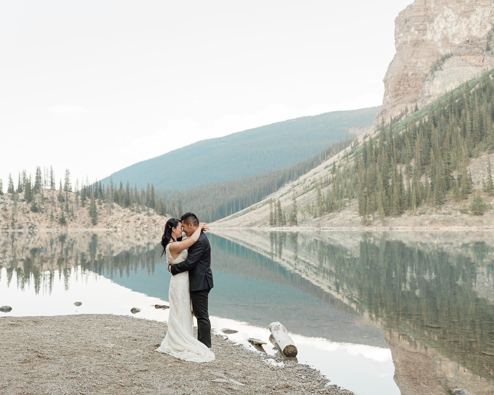 A couple in formal wedding attire embraces near an alpine lake in Banff National Park. 