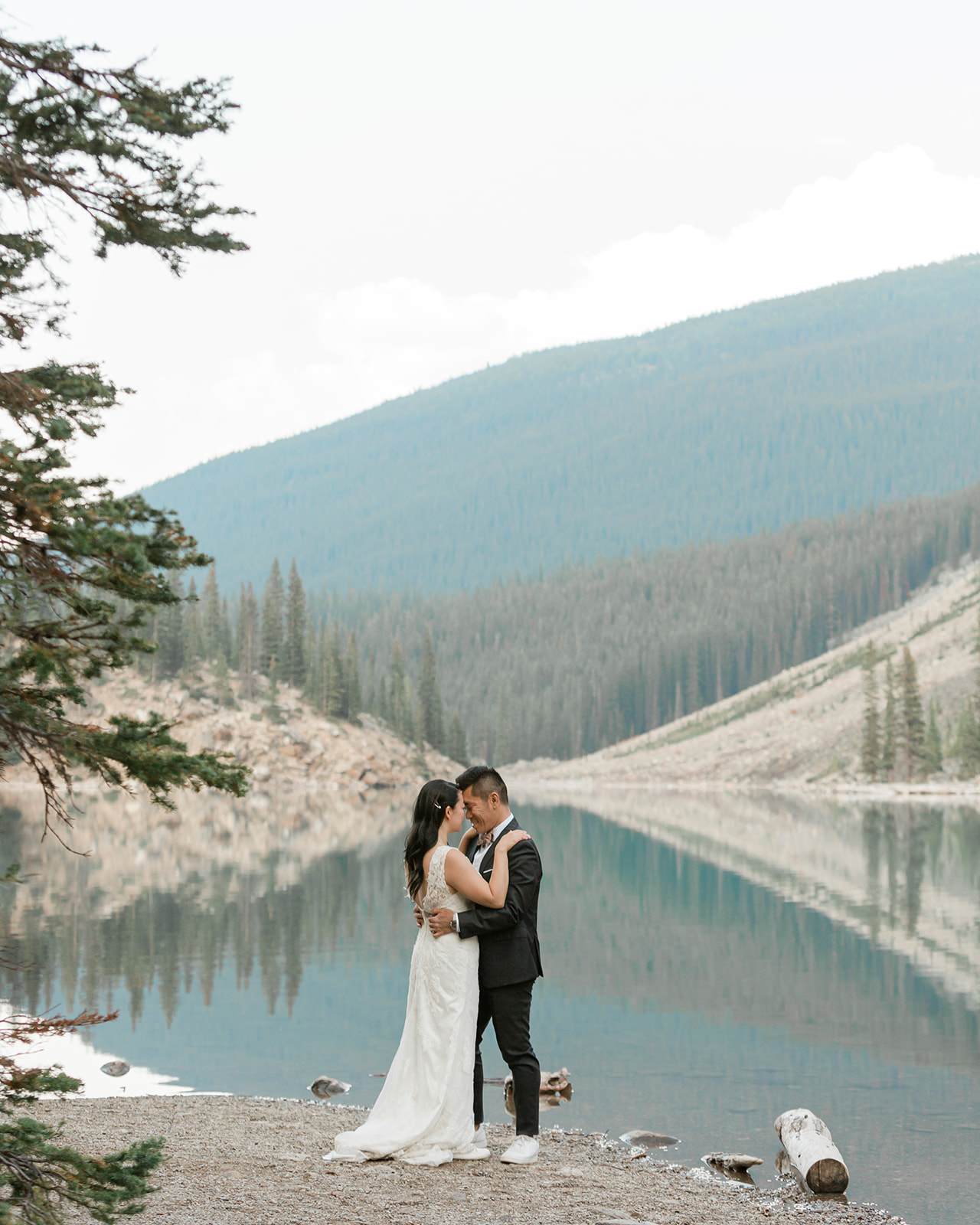 A groom in a black suit holds his bride near an alpine lake during their Banff elopement. 