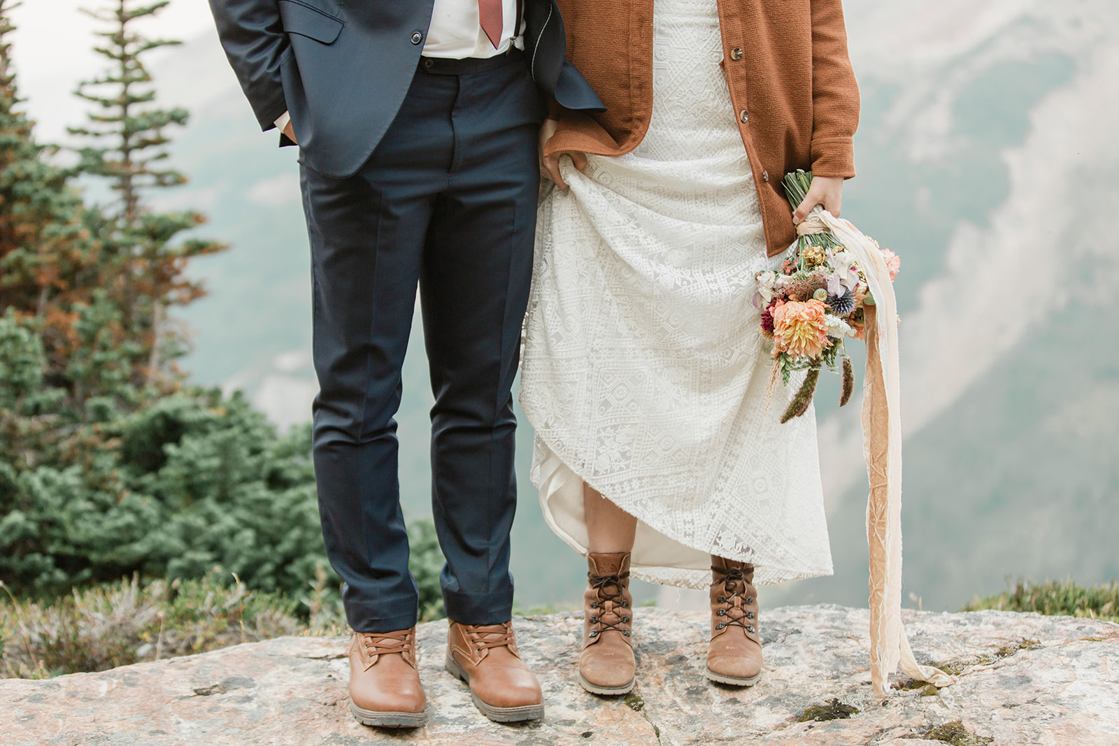 An adventure elopement couple holds hands while standing on a rock while wearing formal wedding attire and hiking boots. 