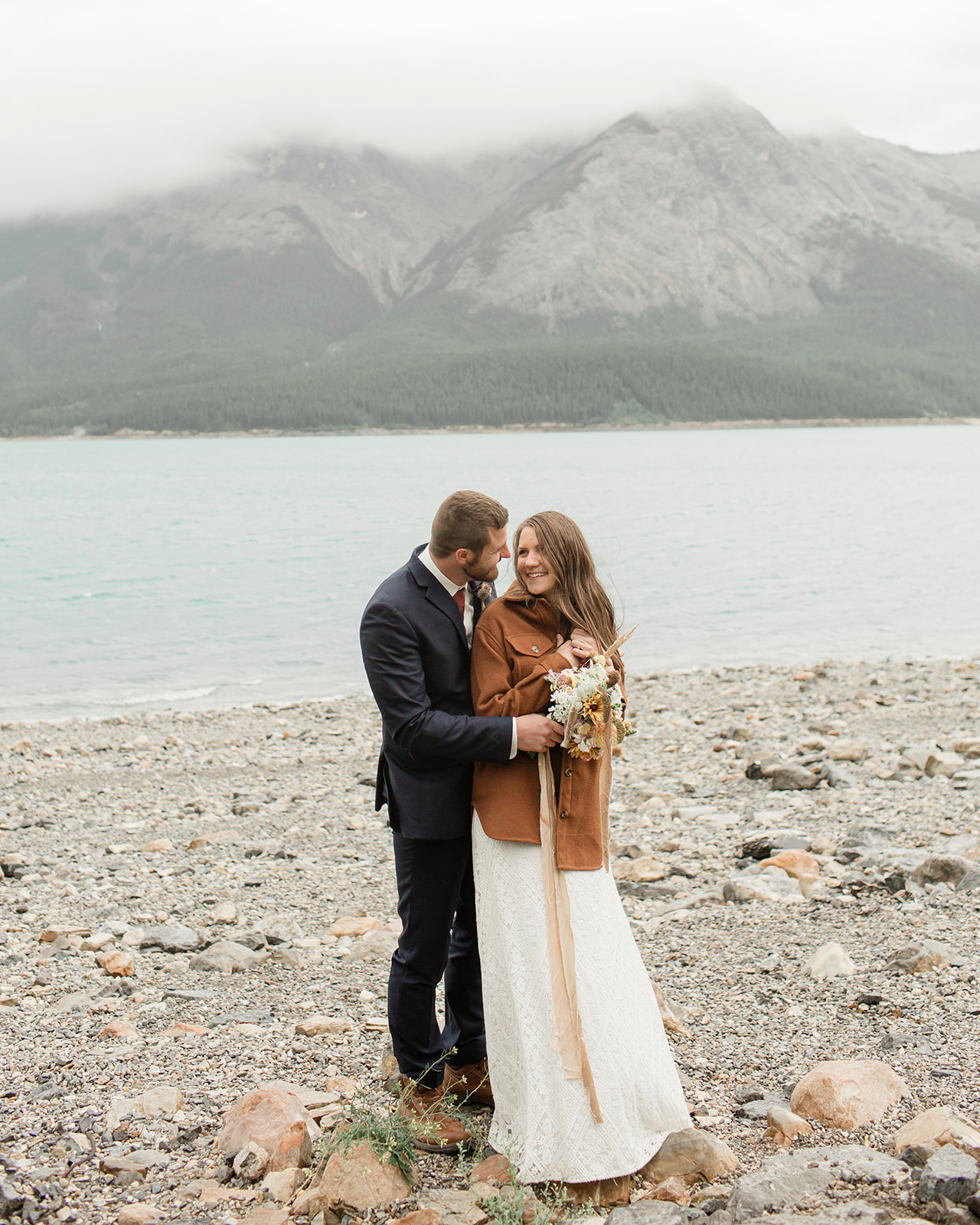 An Alberta wedding couple embraces on a lakeshore near mountains. 