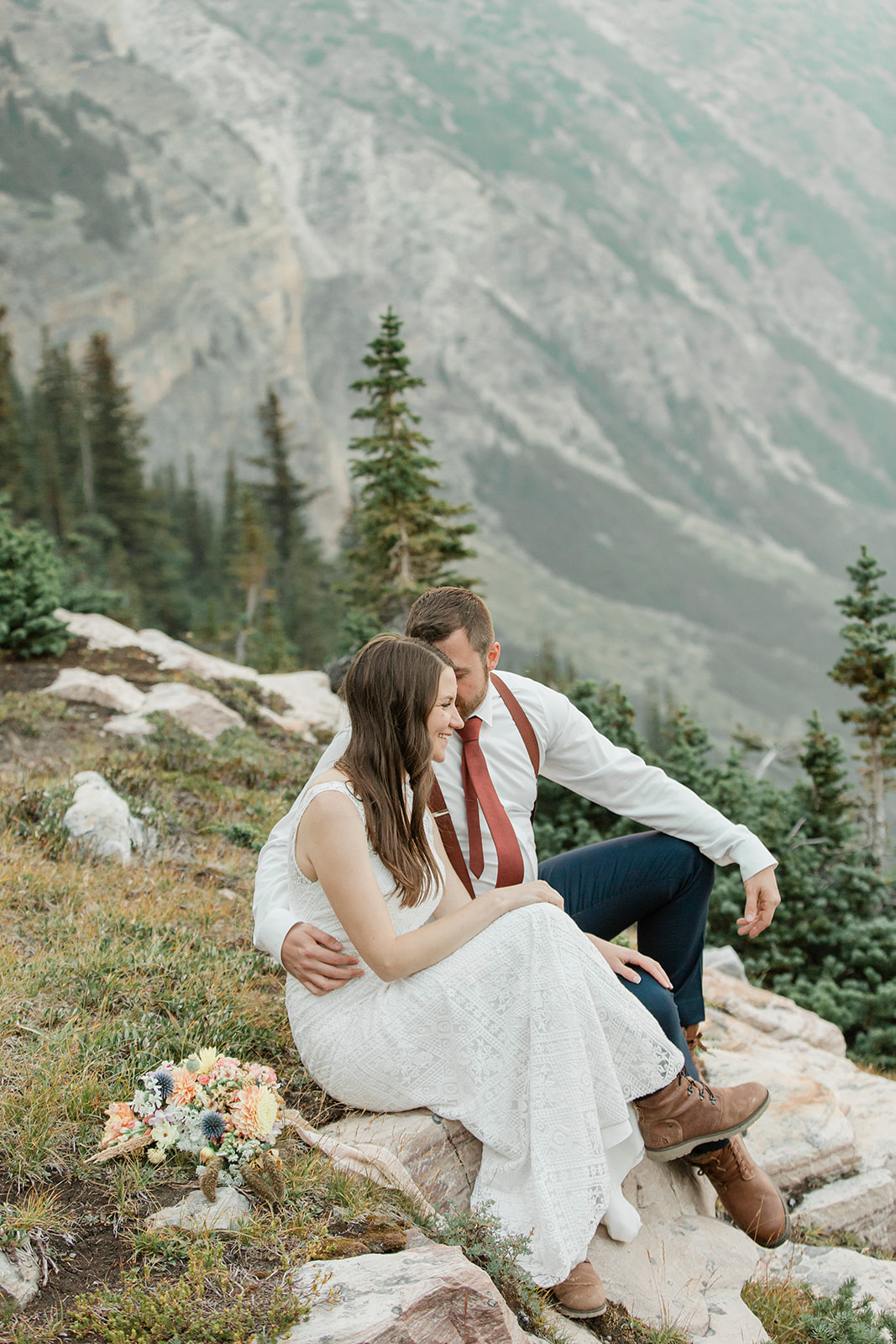 A sustainable elopement couple sits on a rock formation while embracing on an overlook in Alberta. 