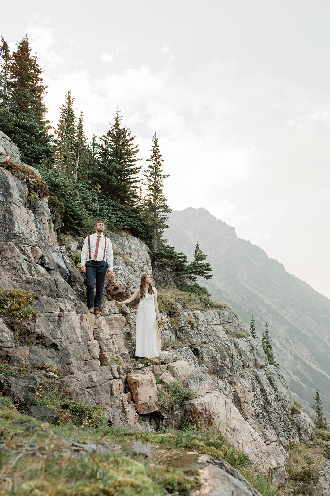 An adventure elopement couple stands on a rock formation while holding hands on an overlook in Alberta. 