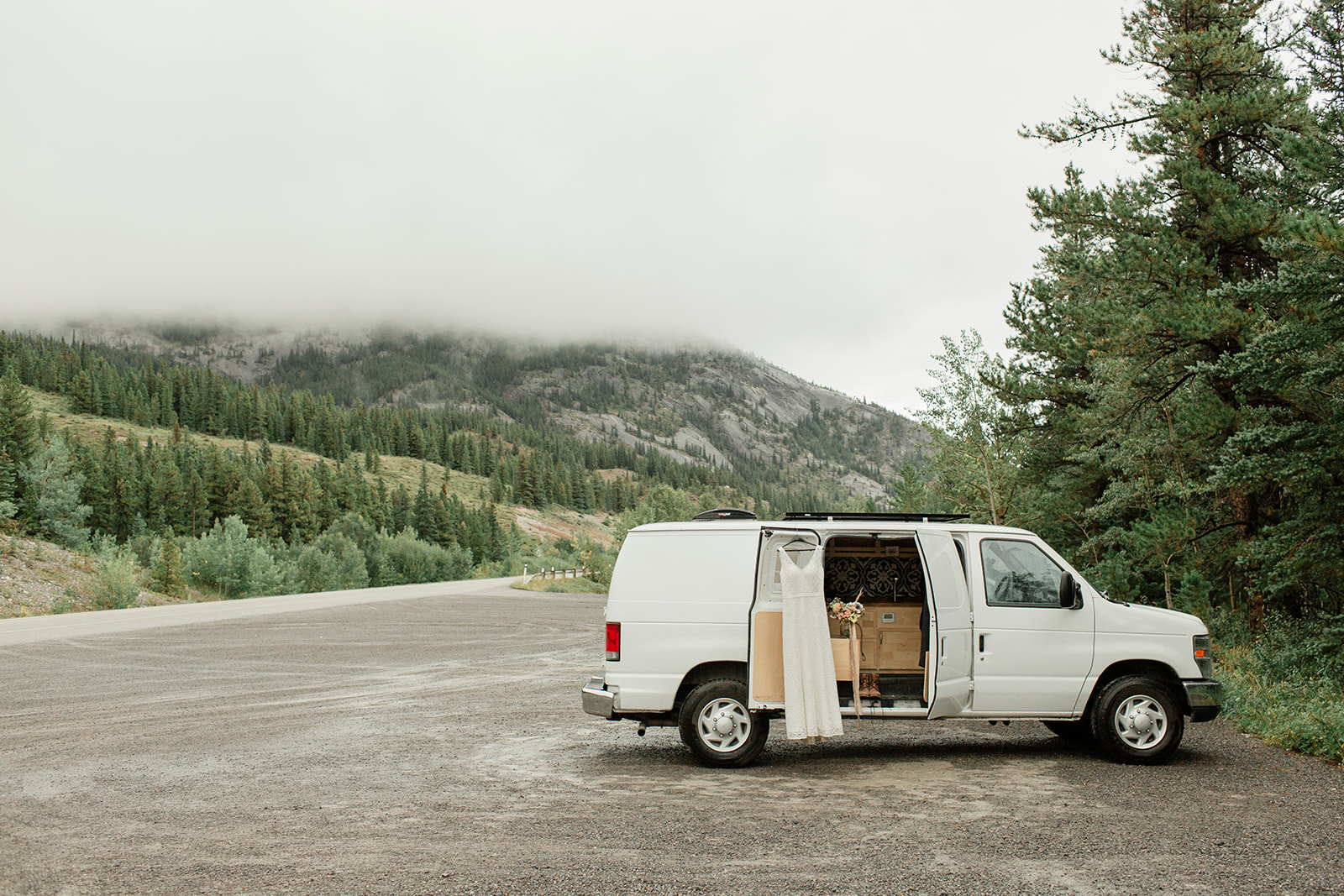 A white van is parked off of a road in Alberta with its door open, displaying a white wedding dress and bridal bouquet. 