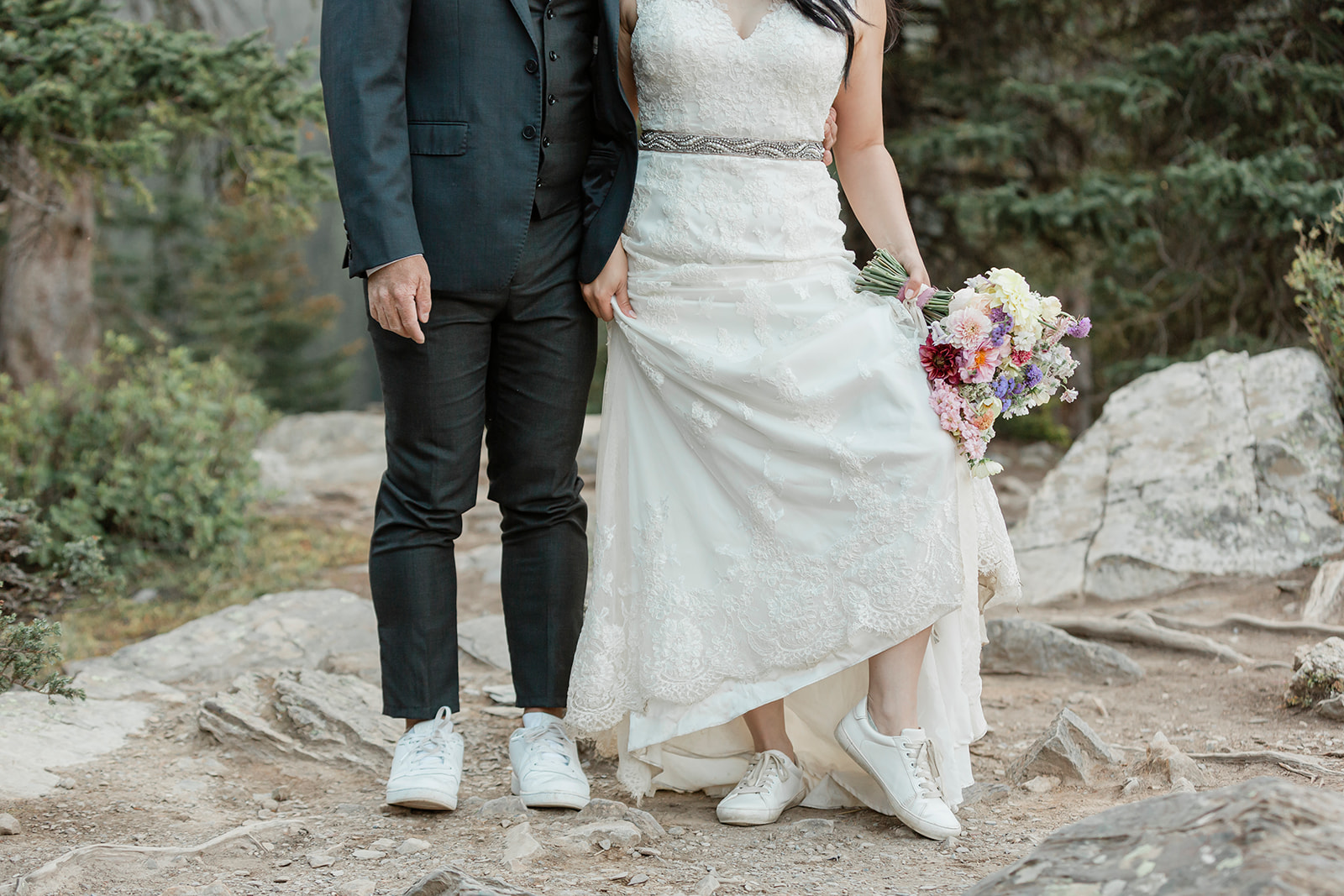 An adventure elopement couple holds hands while standing on a rock while wearing formal wedding attire and white sneakers. 