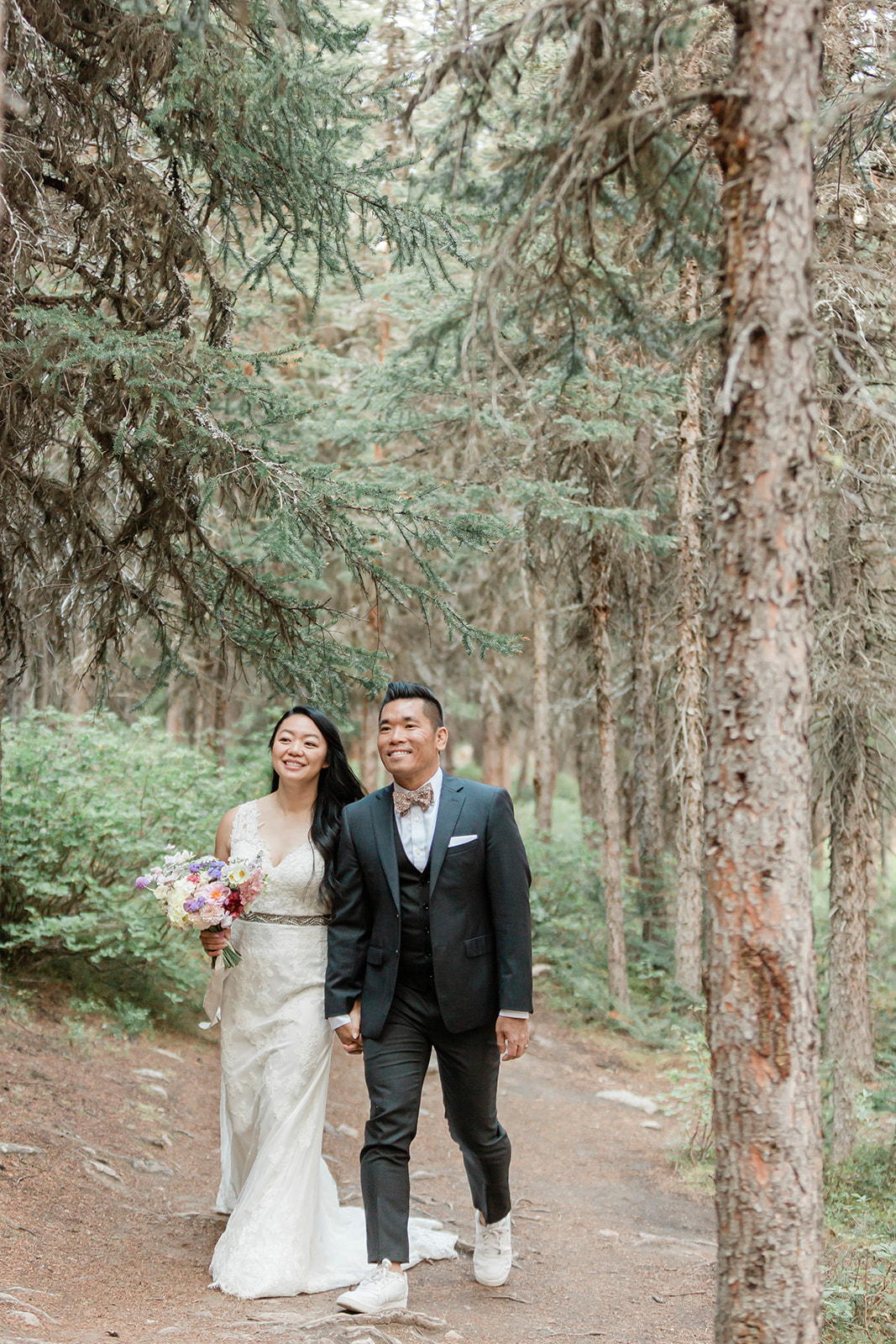 An Alberta elopement couple dressed in formal wedding attire holds hands while walking through a forest.