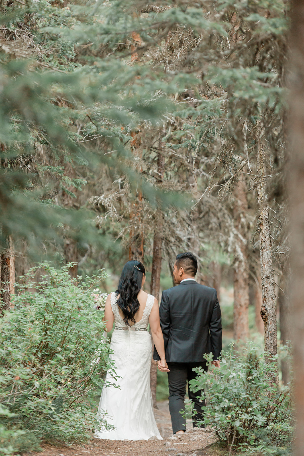 An Alberta elopement couple dressed in formal wedding attire holds hands while walking through a forest.