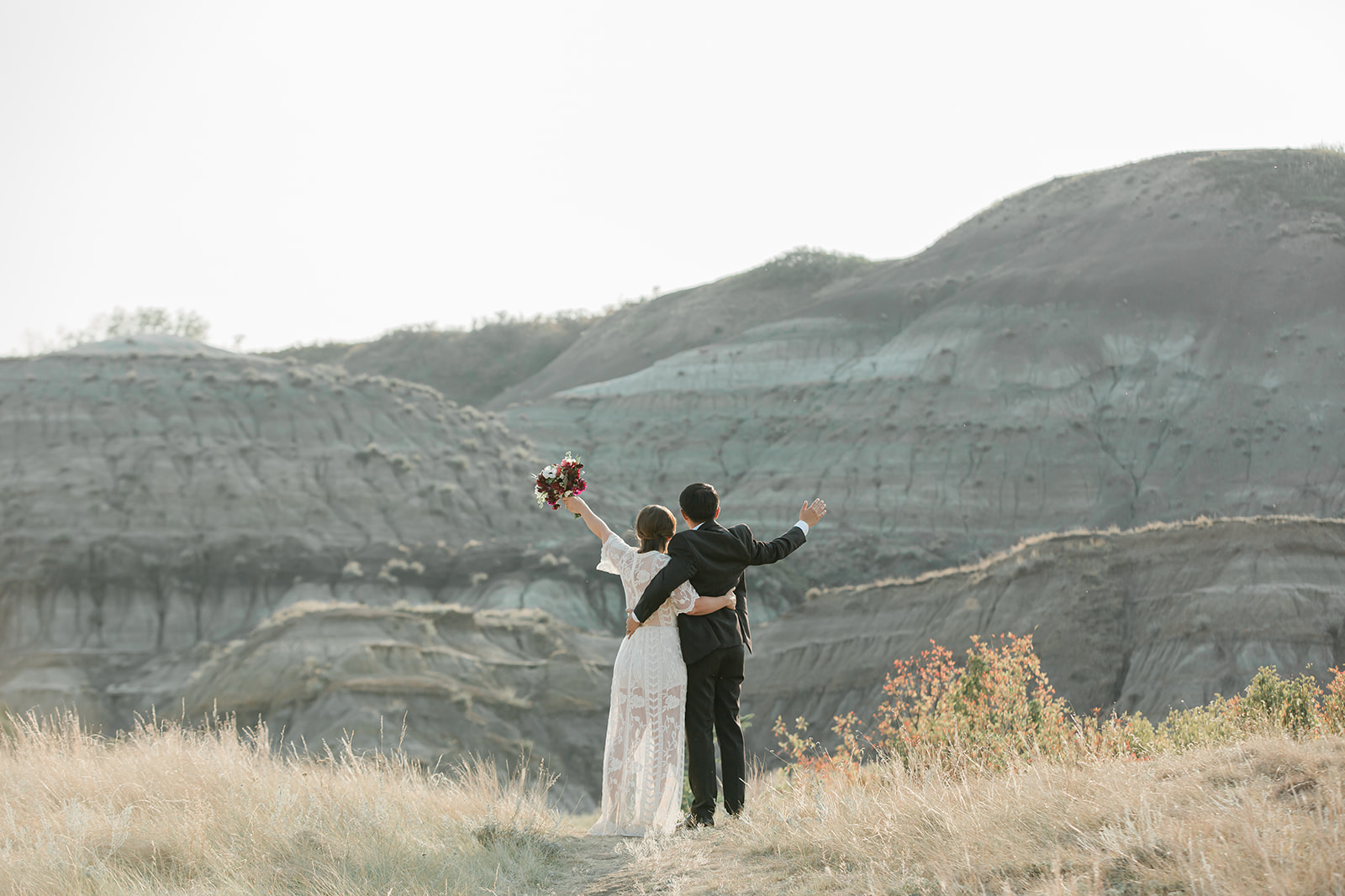 An Alberta elopement couple dressed in formal wedding attire cheers while looking out onto Horsethief Canyon in Alberta, Canada.