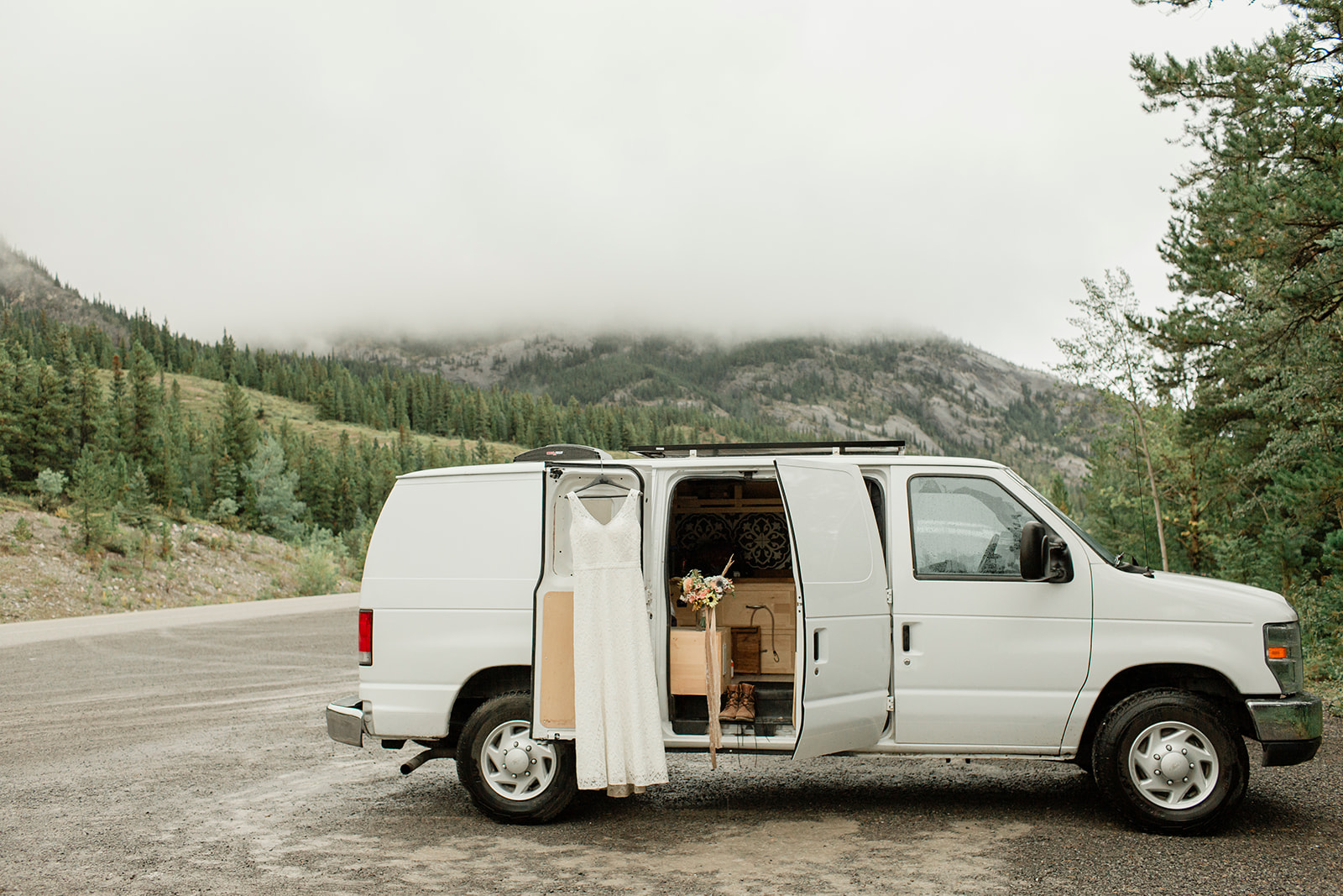 A white van door is parked in Banff National Park for a Banff elopement and it's door open with a wedding dress hanging on it.