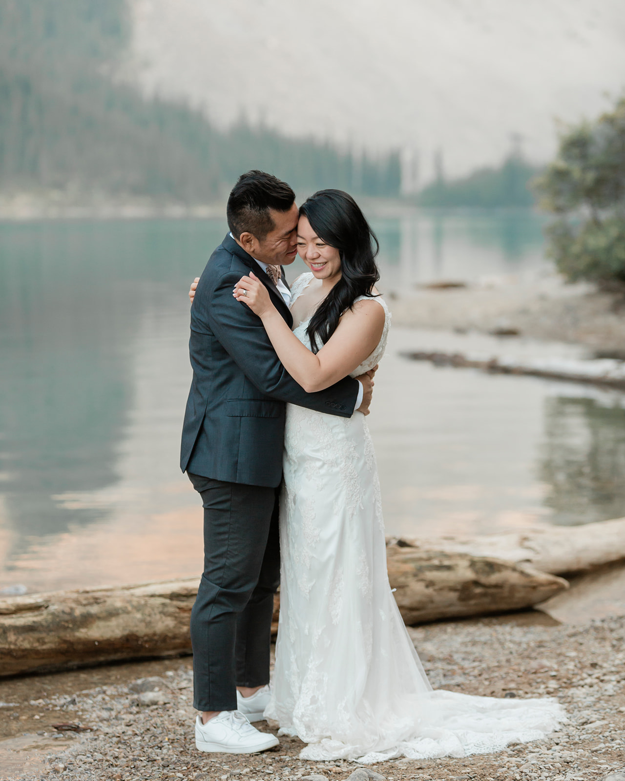 A bride wearing a long, white wedding gown is embraced by her groom while dancing near an alpine lake in Banff National Park.