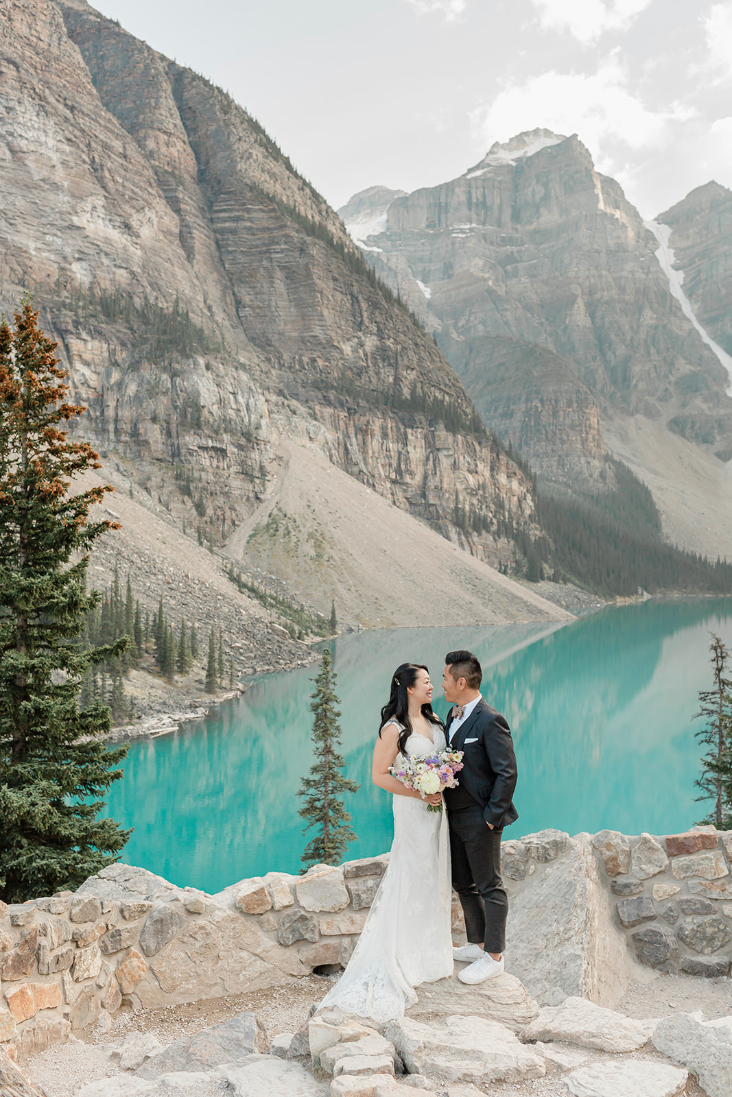 An elopement couple in formal wedding attire stands on an overlook at Moraine Lake in Banff National Park. 